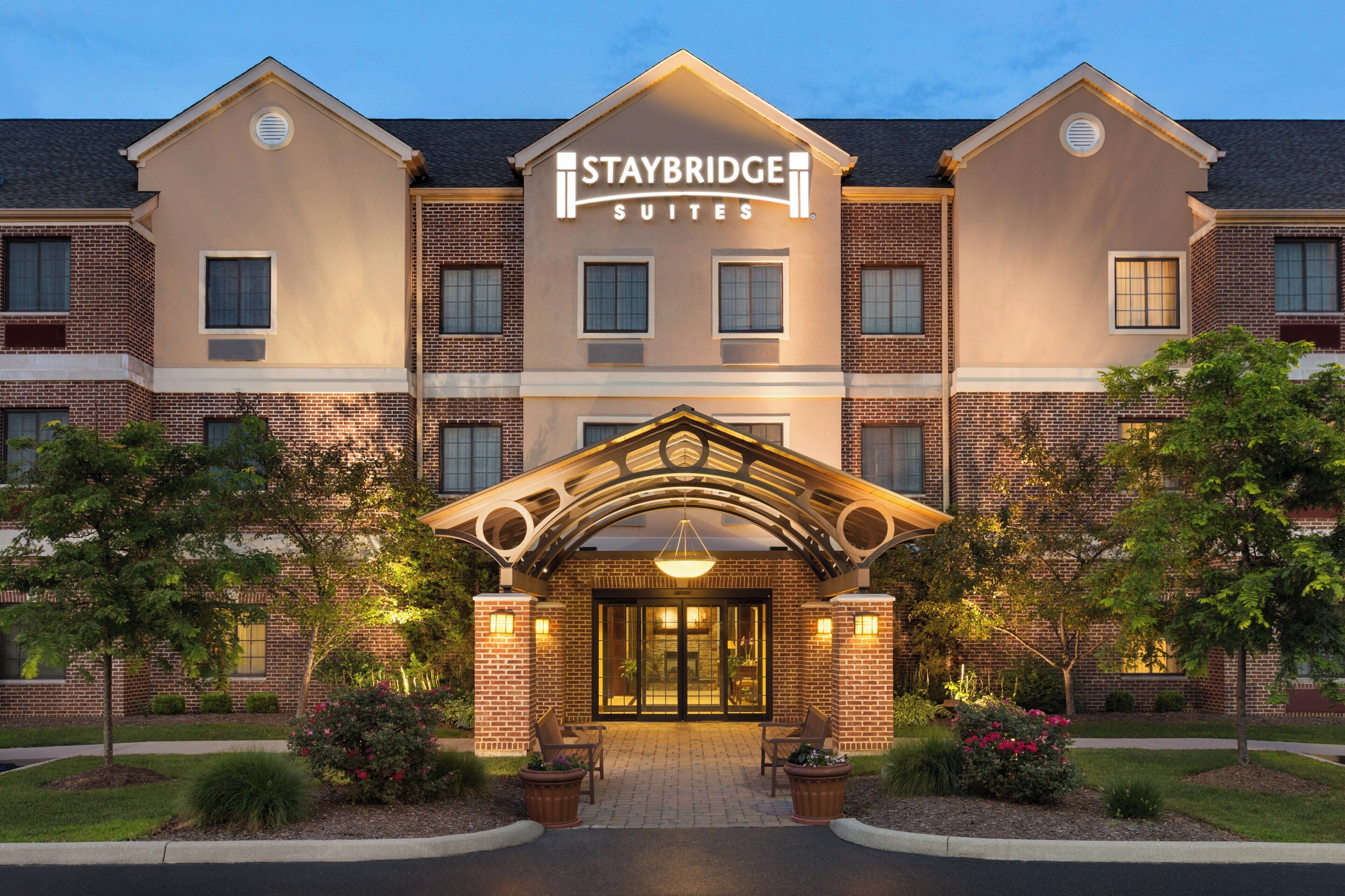 Un hotel Staybridge Suites con fachada de ladrillo, una entrada central con dosel y un camino de entrada ajardinado.