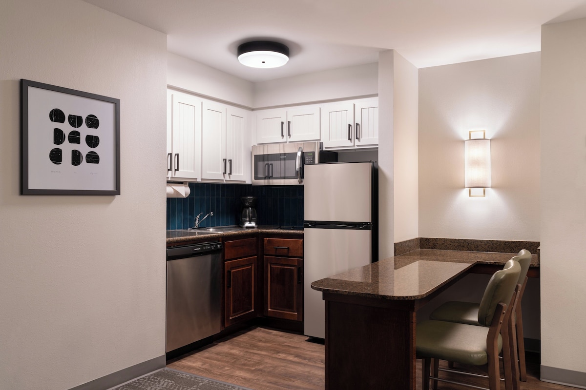 Una cocina moderna con armarios blancos, microondas, refrigerador, cafetera y una barra con sillas.