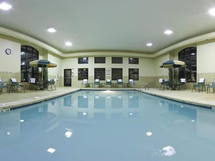 A large indoor swimming pool with multiple lounge chairs and umbrellas, surrounded by windows and a clock on the wall.