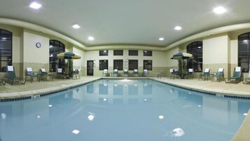 Uma piscina interior ampla com várias espreguiçadeiras e guarda-sóis, cercada por janelas e um relógio na parede.
