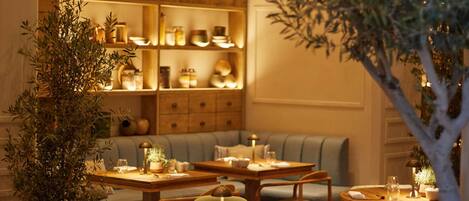 A restaurant with multiple round tables and chairs, a variety of kitchenware displayed on shelves, and a potted tree in the foreground.