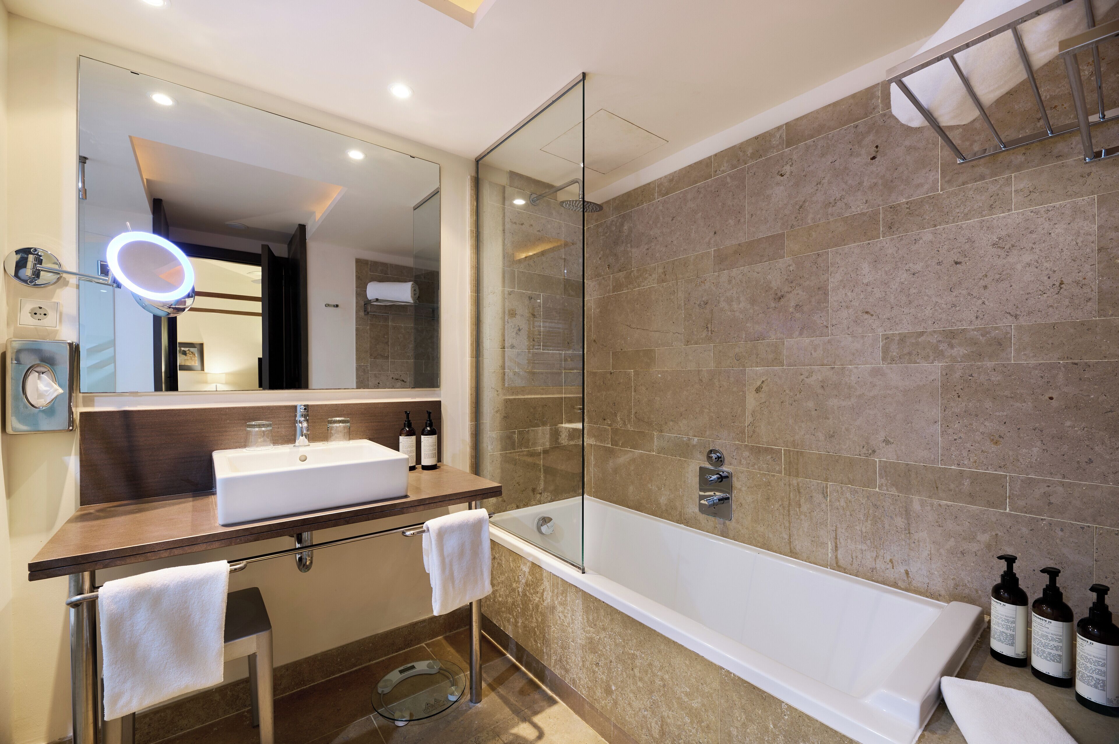 Un bagno moderno con doccia vetro, uno specchio grande illuminato da una luce ad anello, una vasca da bagno bianca e un mobile lavabo con lavandino e due flaconi di shampoo.