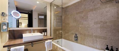 A modern bathroom with a glass-enclosed shower, a large mirror with a ring light, a white bathtub, and a vanity with a sink and two bottles of shampoo.