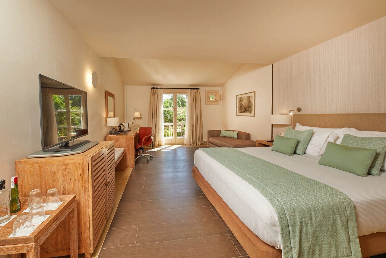 Habitación de hotel con cama, televisor, escritorio con computadora, silla y vistas al exterior.