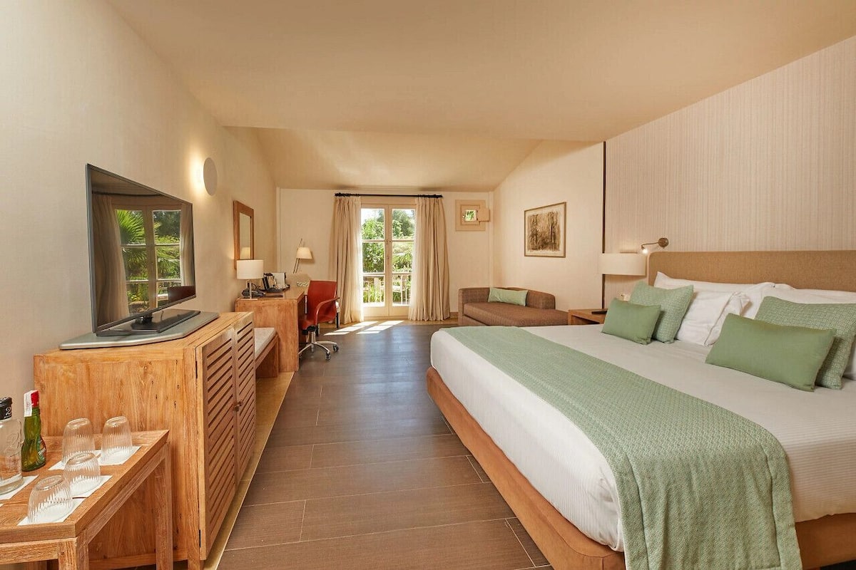 Habitación de hotel con cama, televisor, escritorio con computadora, silla y vistas al exterior.