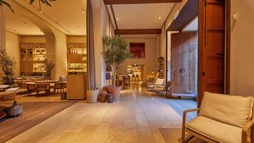 Un hall d’entrée spacieux avec un plafond haut, des poutres en bois et un espace salon central. On y trouve plusieurs tables et chaises, ainsi qu’une grande plante en pot au milieu.