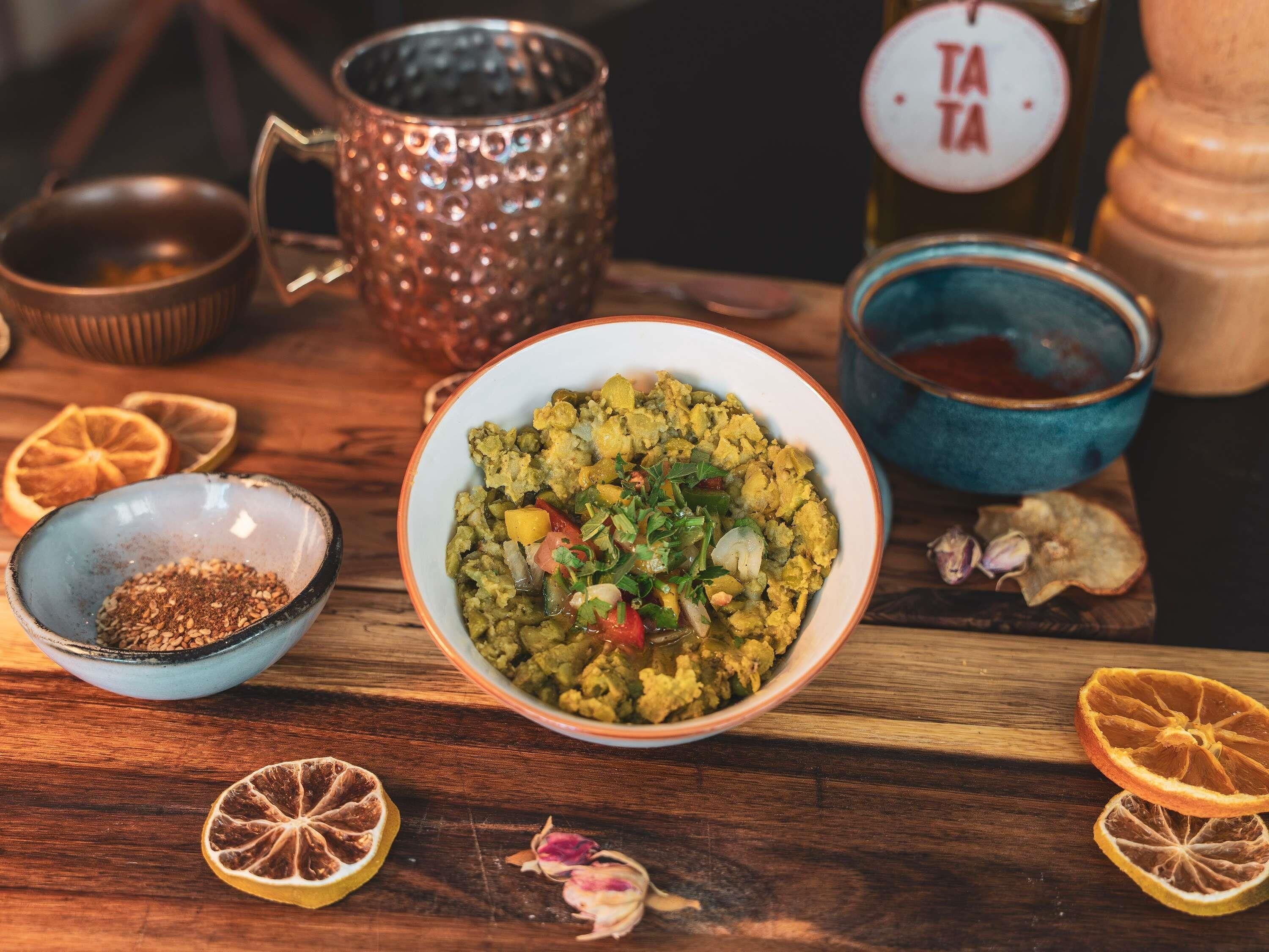 A bowl of food on a wooden table with a copper mug, a blue bowl, and a bottle labeled 'TATA' in the background.