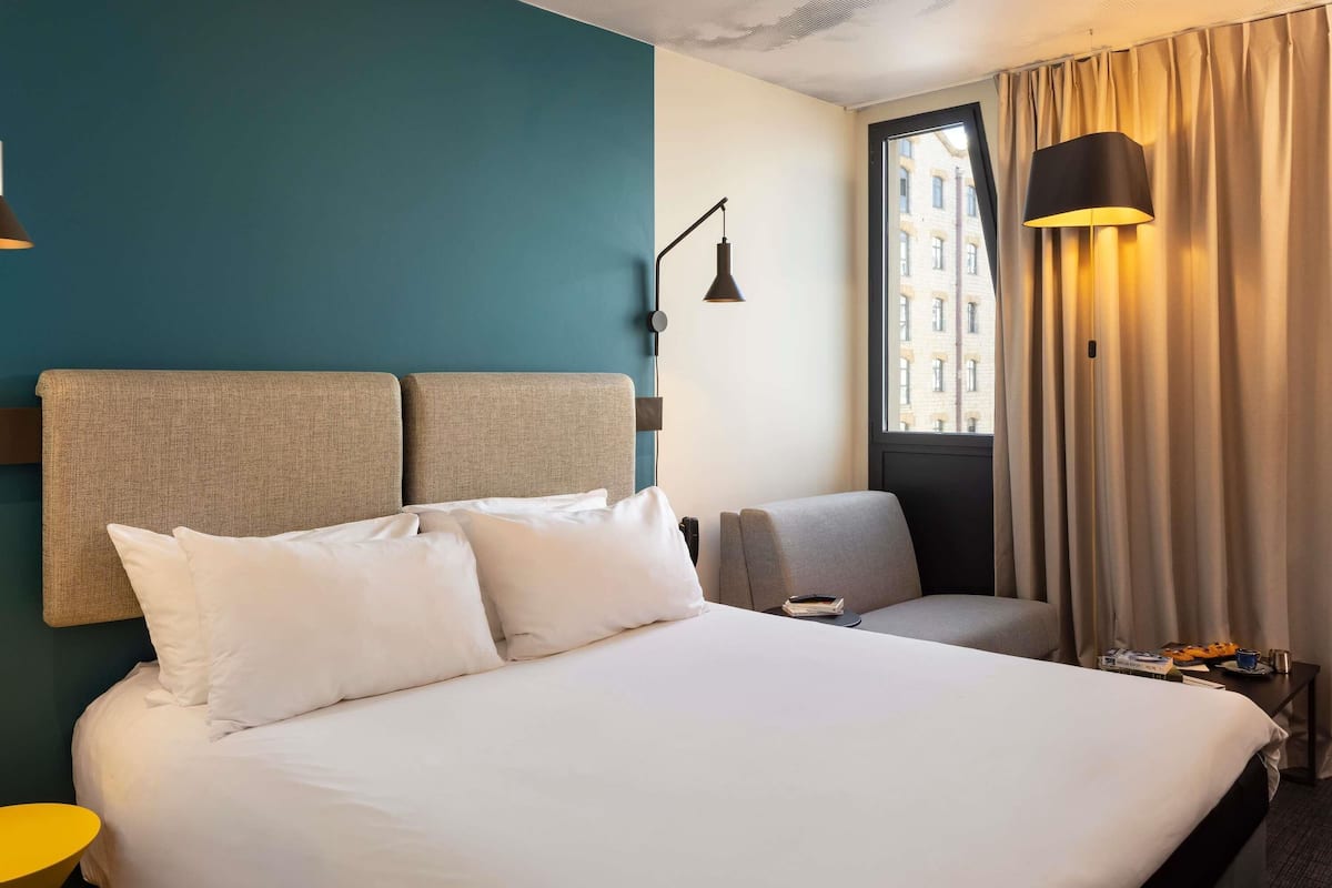 A hotel room with a large bed, a chair, a desk, a lamp, and a window with curtains.