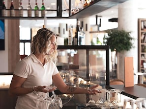 A bar area with shelves of bottles and glasses, a counter with a person serving drinks, and a plant in the background. - Ibis Marseille Centre Euromed (Marseille)