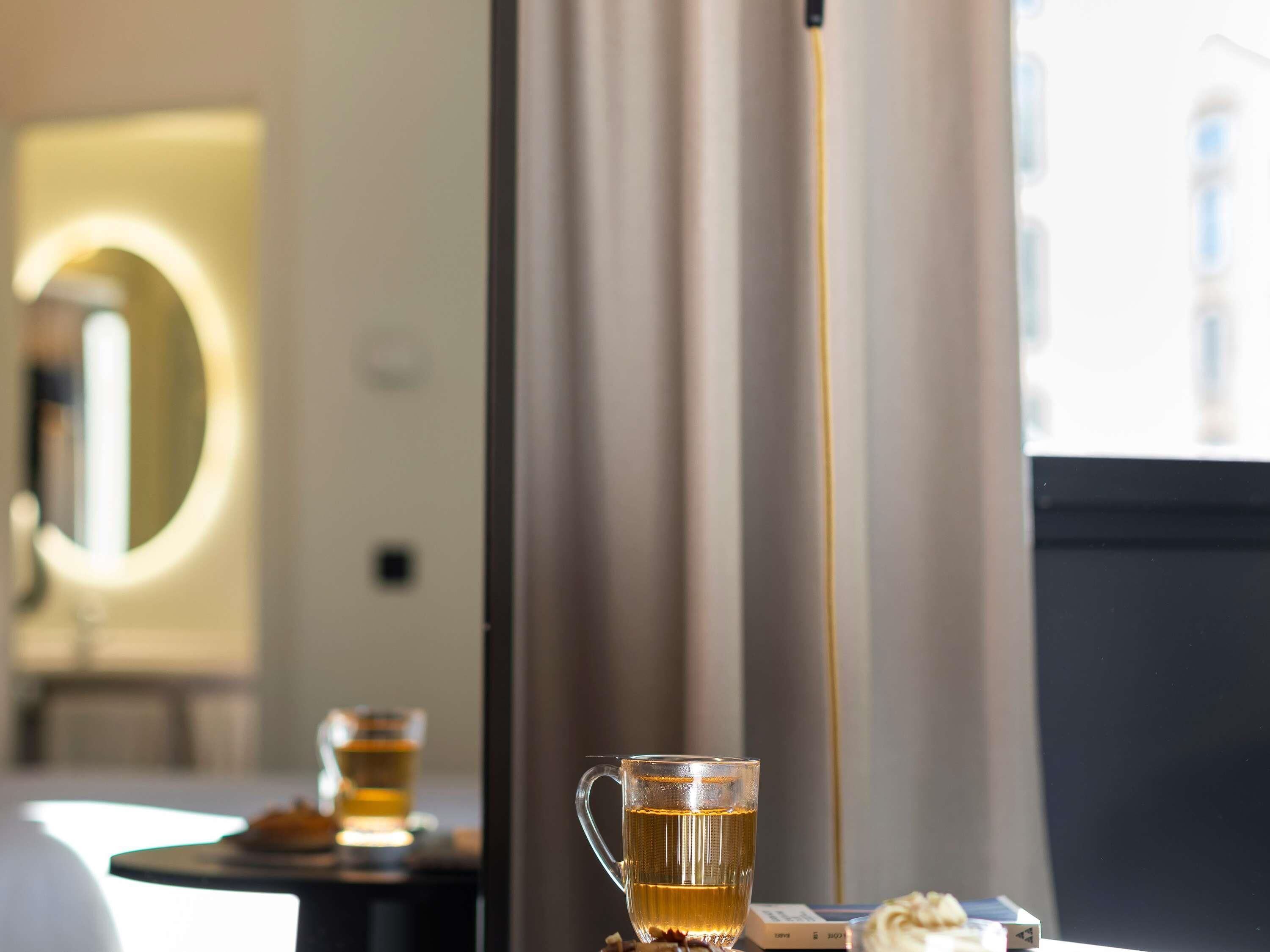 A hotel room with a bed, a bedside table with a cup of tea, a mirror, and a window with curtains.