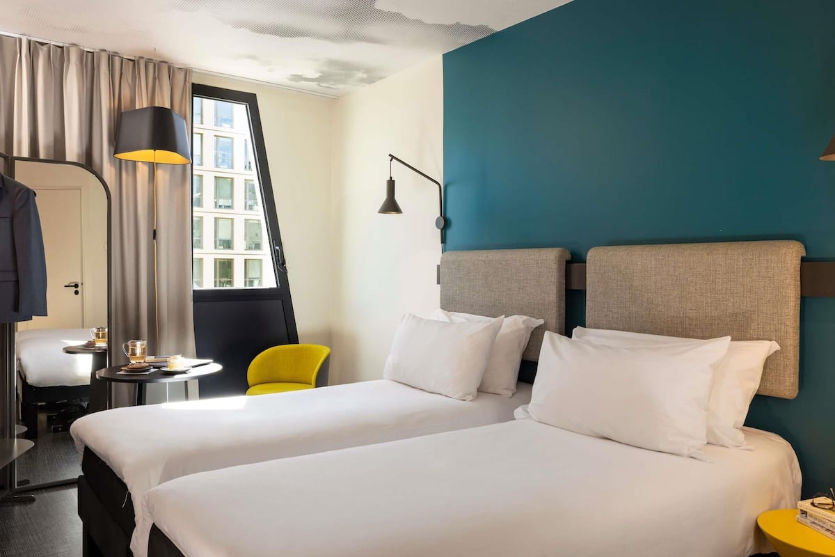 A hotel room with two beds, a desk, a chair, a lamp, and a view of buildings through a window.