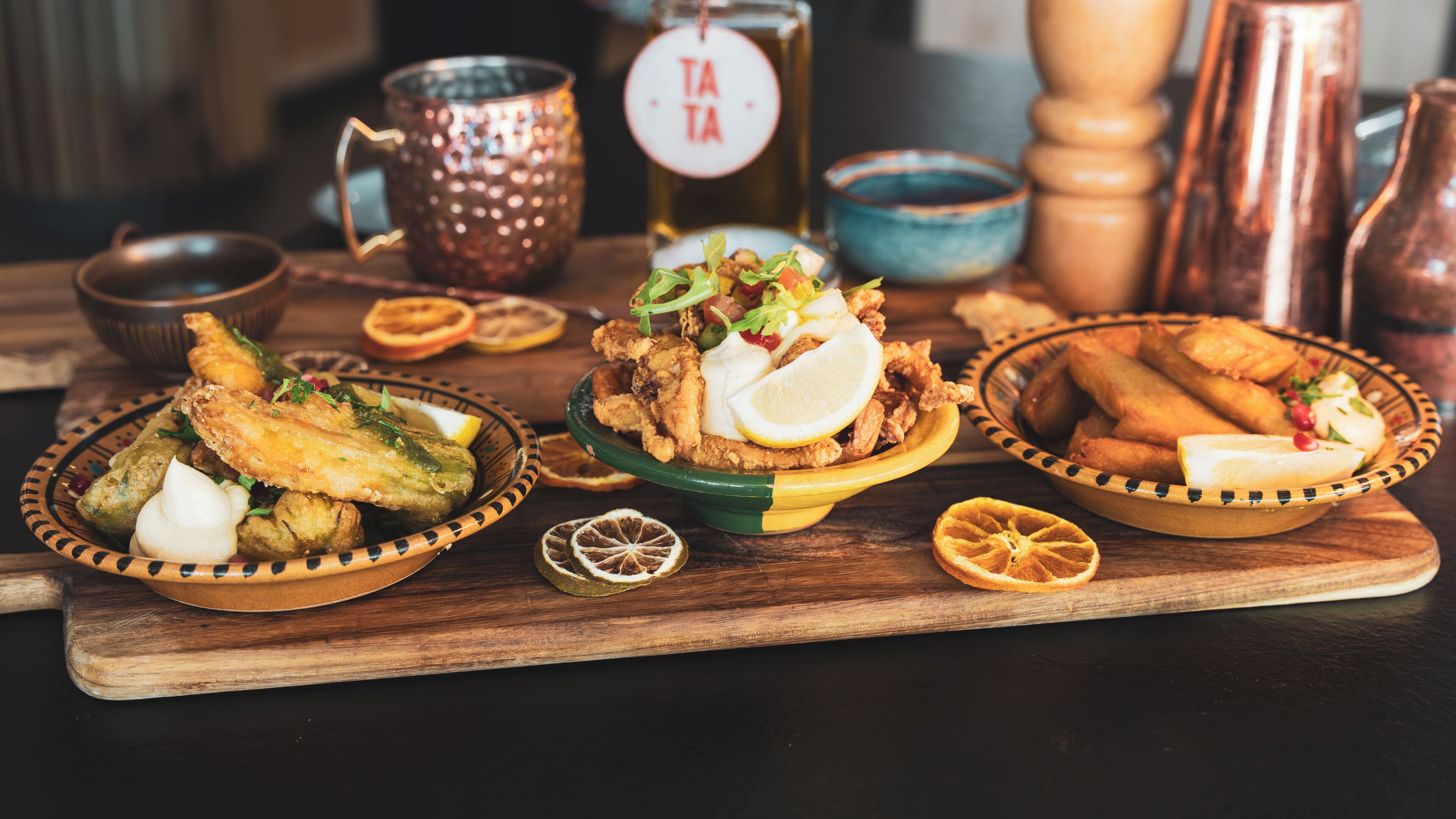 A wooden table with three bowls of fried food, a copper mug, a bottle with a label, and a salt shaker.
