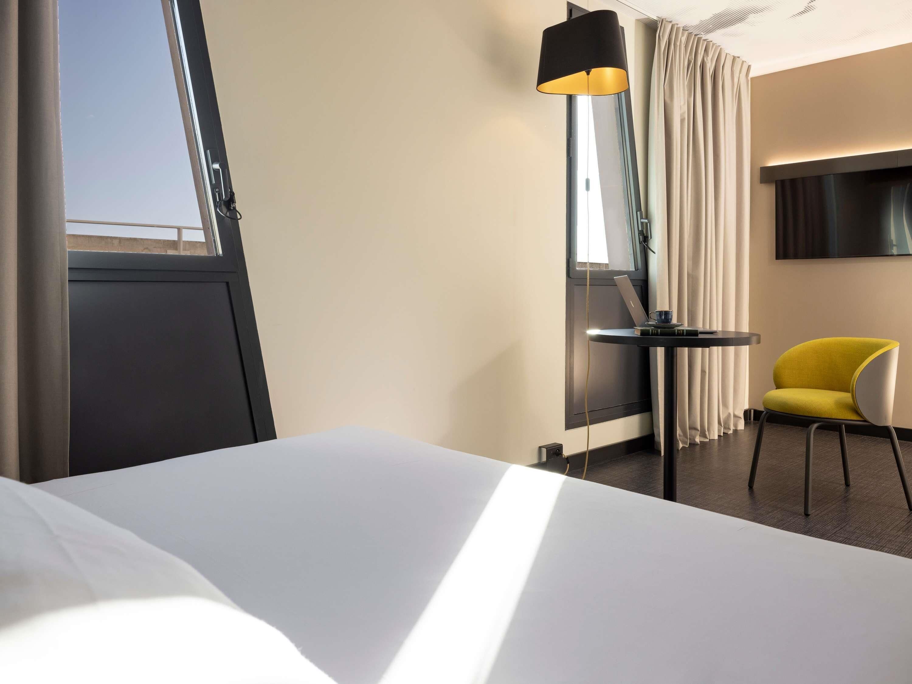 Ein Hotelzimmer mit einem Bett, einem Schreibtisch mit einem Laptop, einem gelben Sessel, einer Stehlampe und einem an der Wand montierten Fernseher.