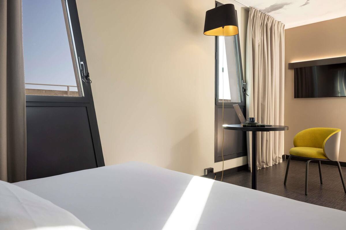 A hotel room with a bed, a desk with a laptop, a yellow chair, a standing lamp, and a TV mounted on the wall.