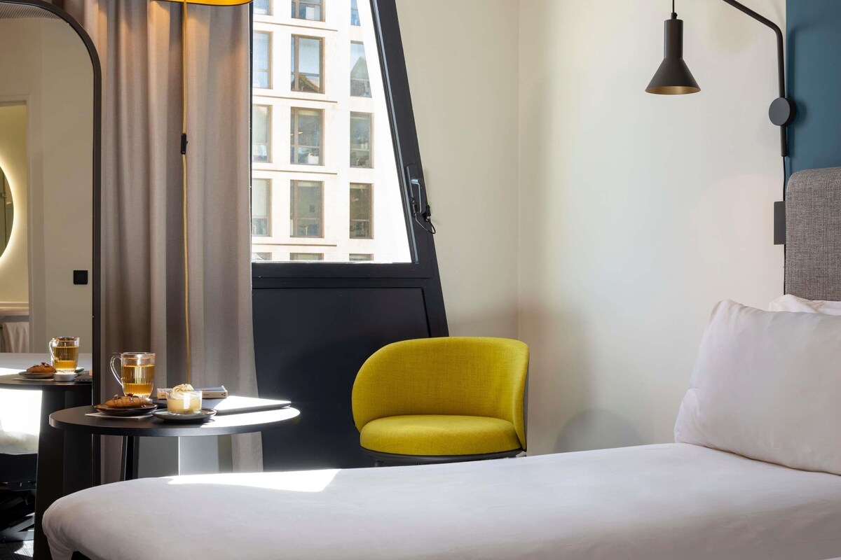 Una habitación de hotel con una cama, una silla amarilla, una mesa redonda con una taza de té y una ventana con cortinas.