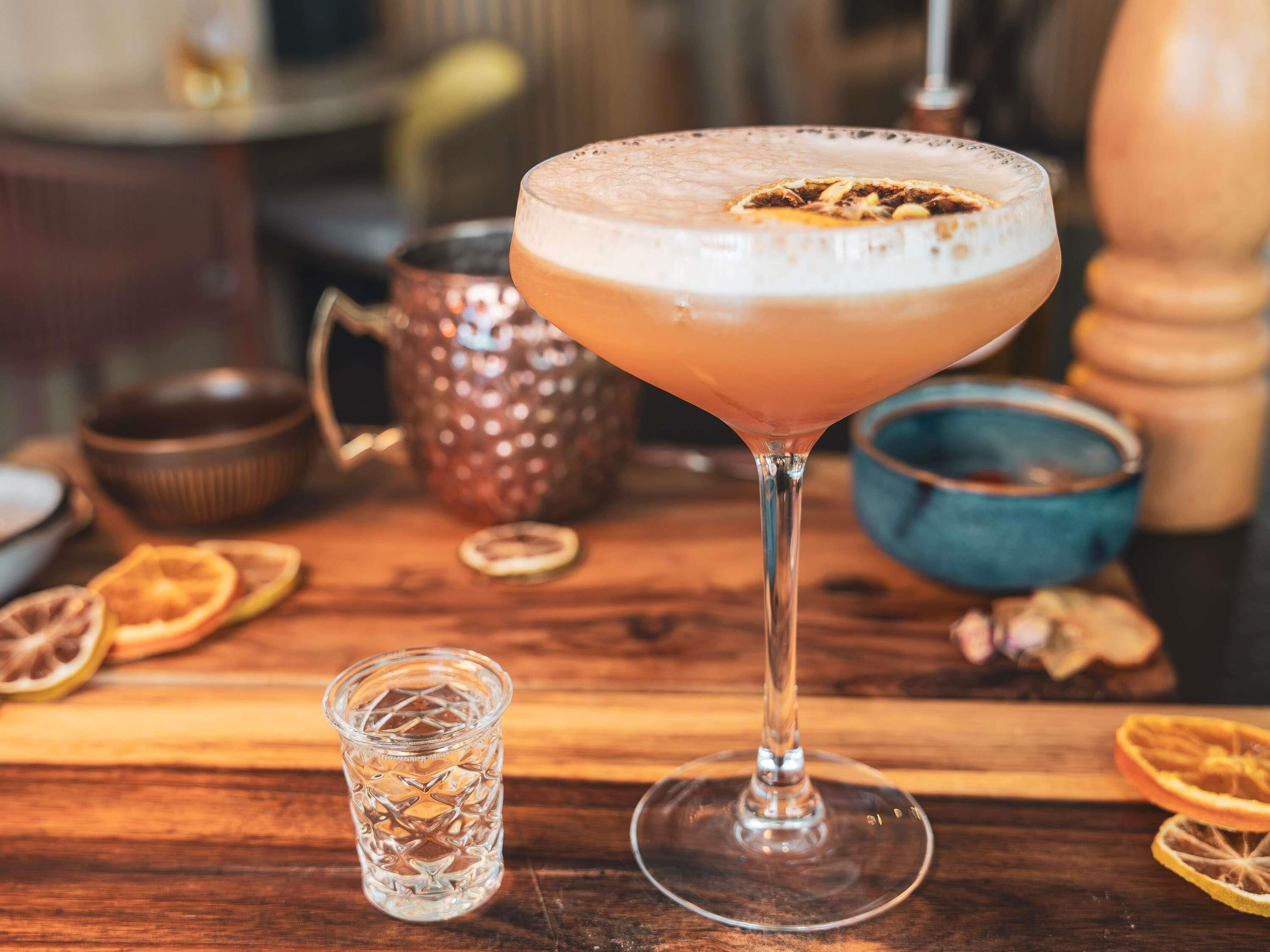 A cocktail in a martini glass on a wooden table with a small glass and dried orange slices.