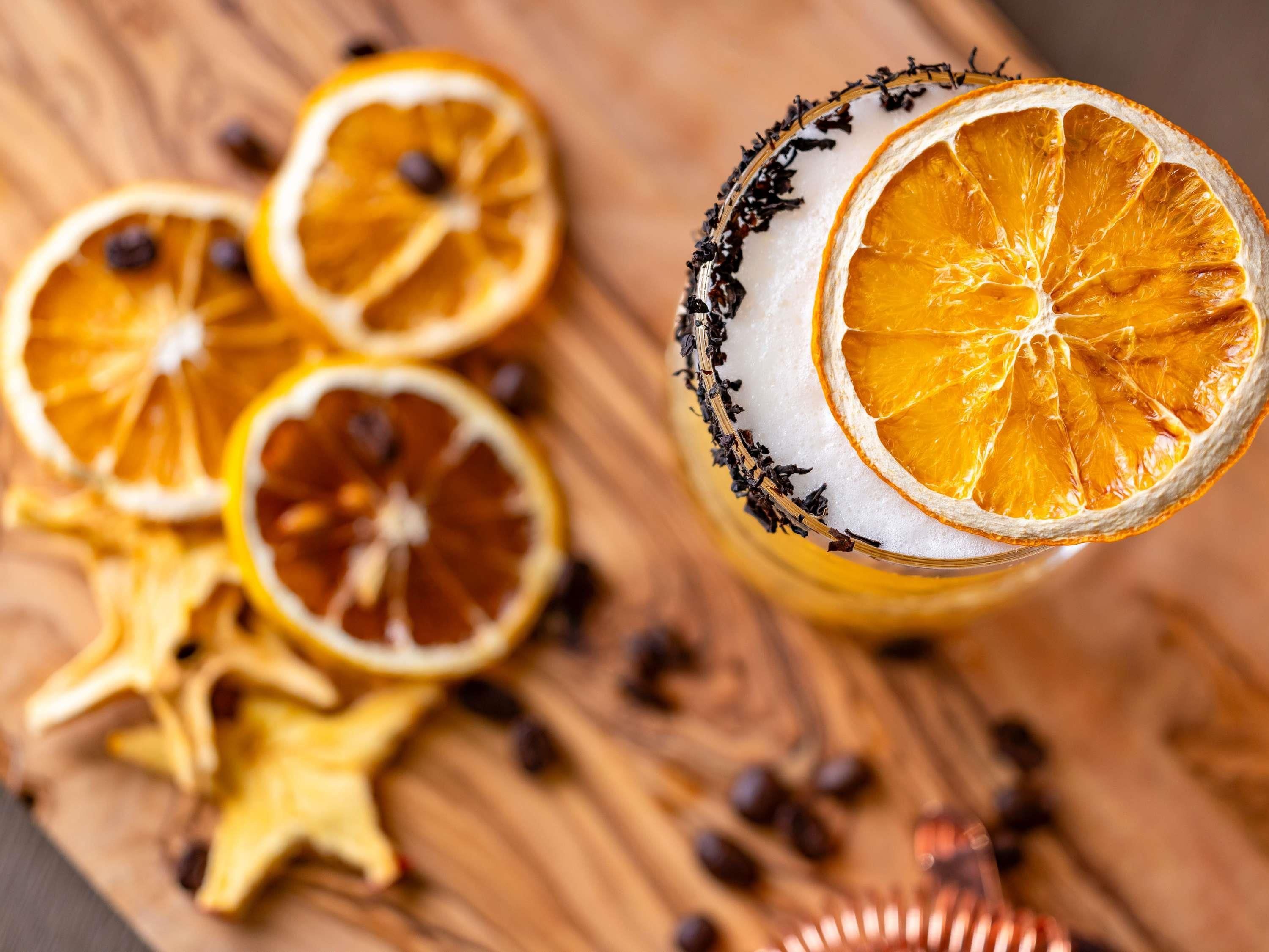 Ein Glas mit einem Rand, der mit getrockneten Orangenscheiben und Kräutern verziert ist, umgeben von weiteren getrockneten Orangenscheiben und sternförmigen Dekorationen auf einer Holzoberfläche.