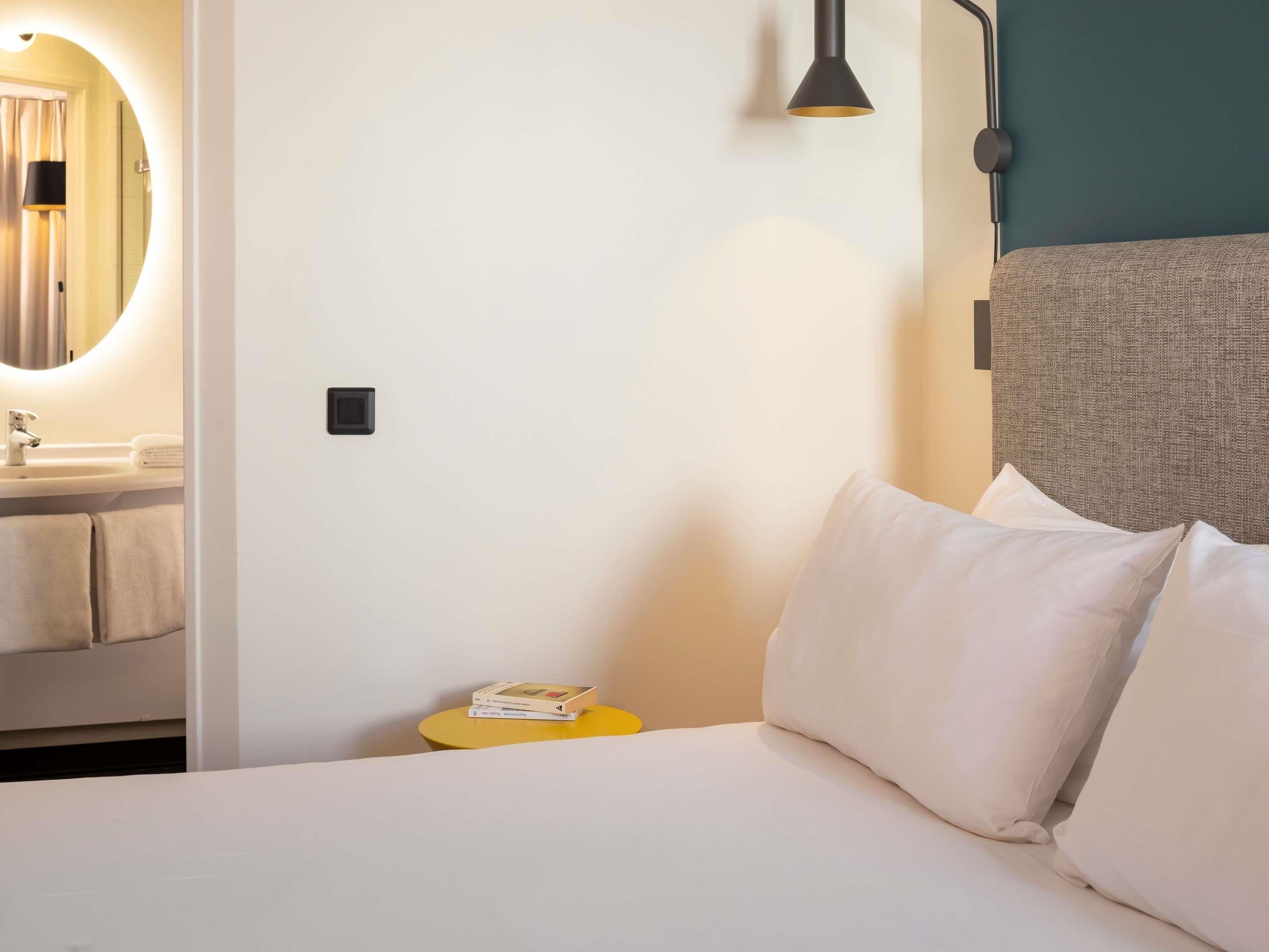 Habitación de hotel con cama, mesita de noche con un libro, lámpara de pared y baño visible a través de una puerta abierta.