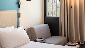 A hotel room with a bed, a grey armchair, a small table with books and a remote control, a lamp, and a window with curtains. - Ibis Marseille Centre Euromed (Marseille)