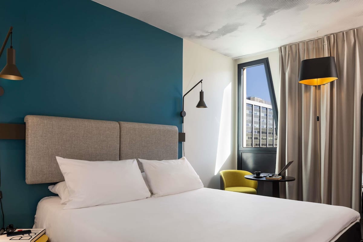 A hotel room with a large bed, a small table with a chair, a window with curtains, and a view of a building outside.