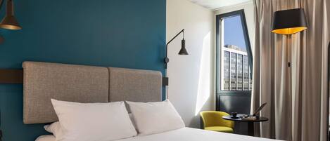 A hotel room with a bed, a yellow chair, a small table, and a window with curtains.