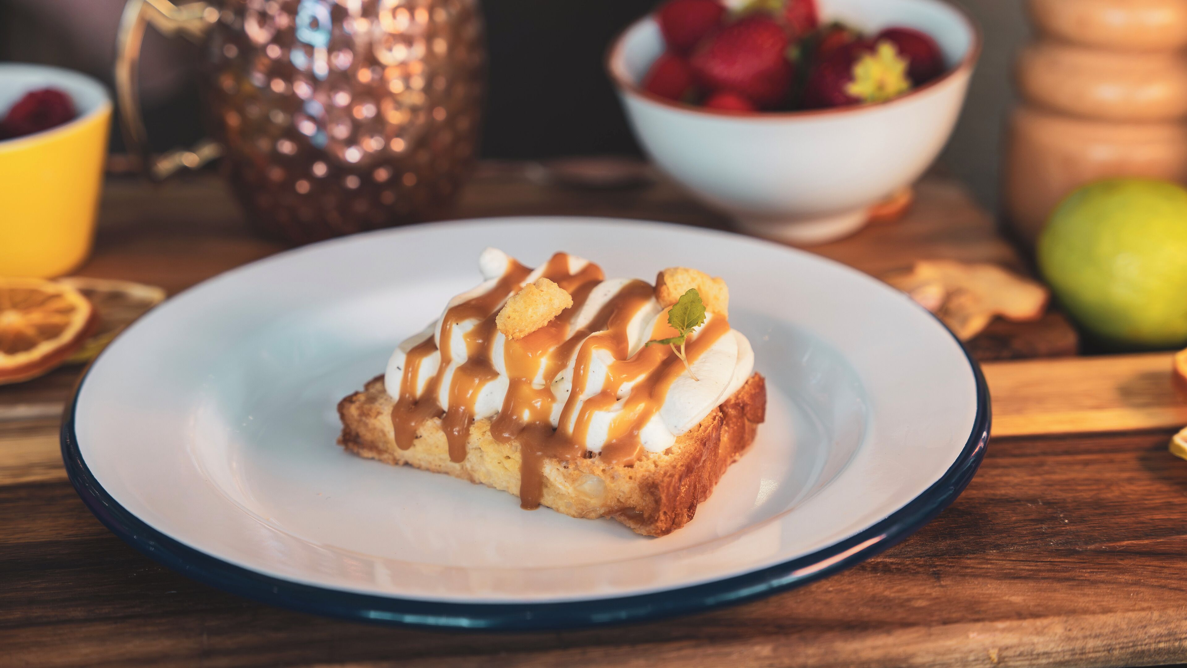 A dessert on a plate with a caramel drizzle, surrounded by fruits and a copper pot.