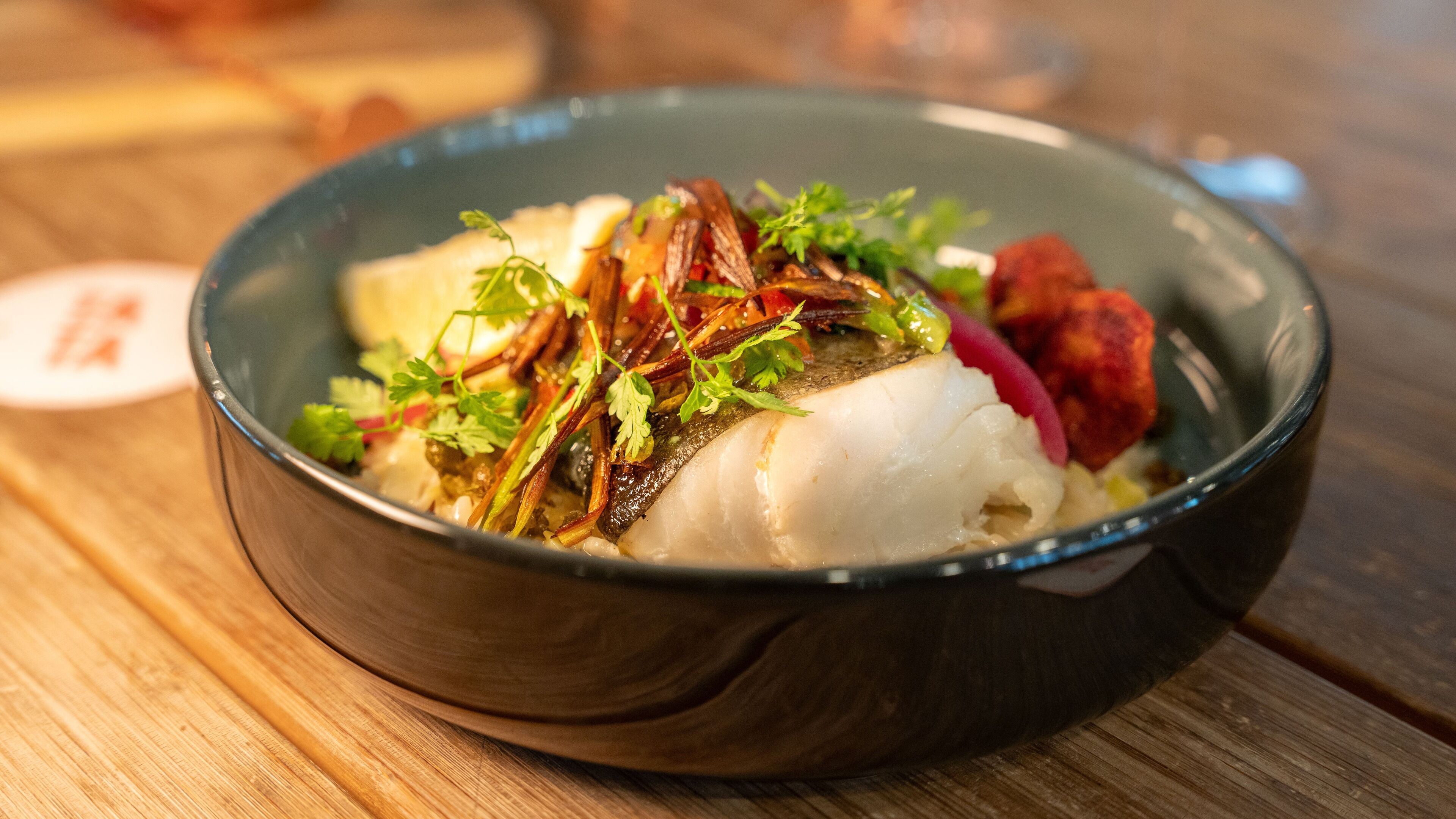 A bowl of food with fish, vegetables, and herbs on a wooden table.