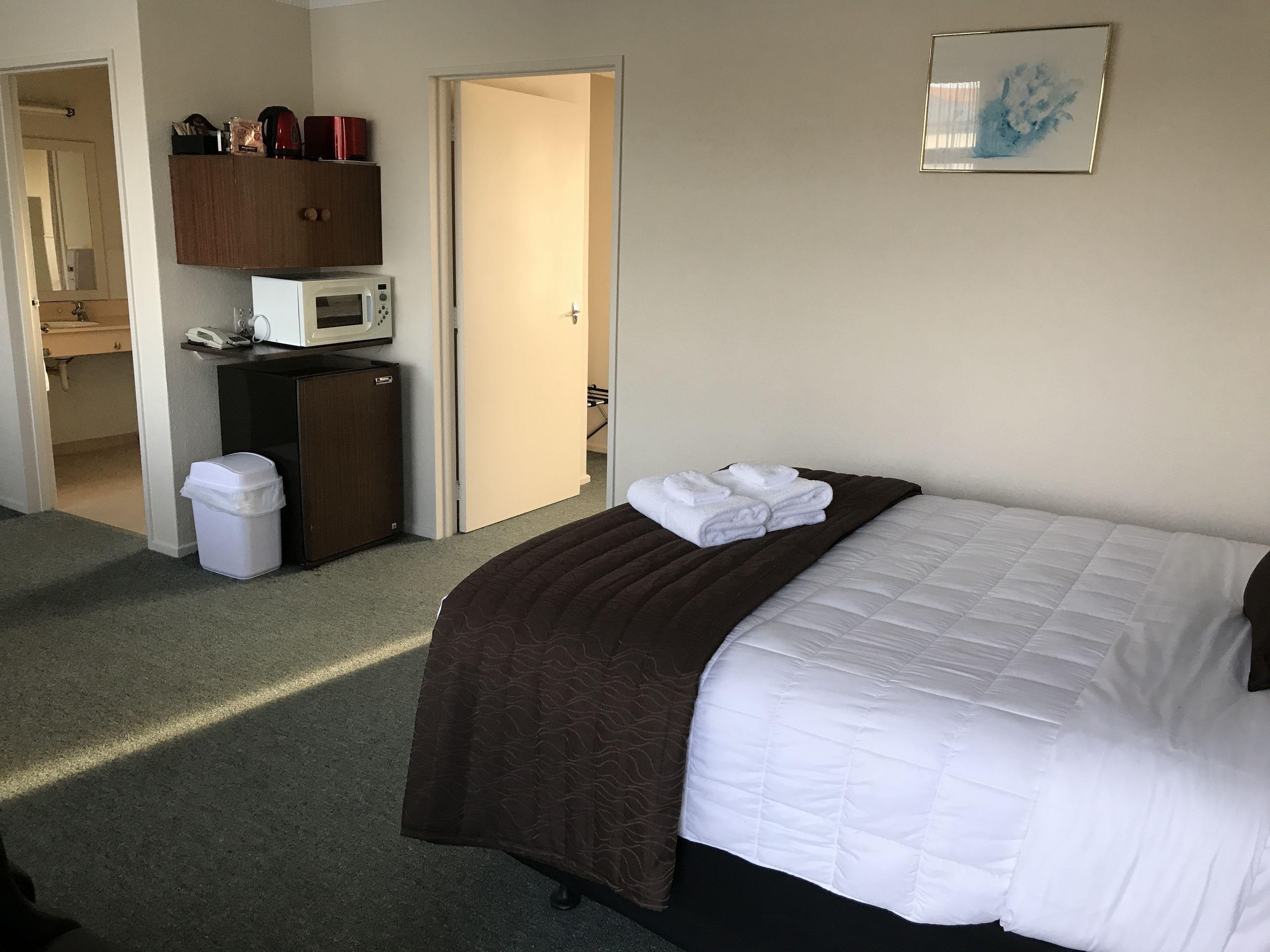 A hotel room with a bed, a microwave, a trash bin, a cabinet, and a door leading to another room.