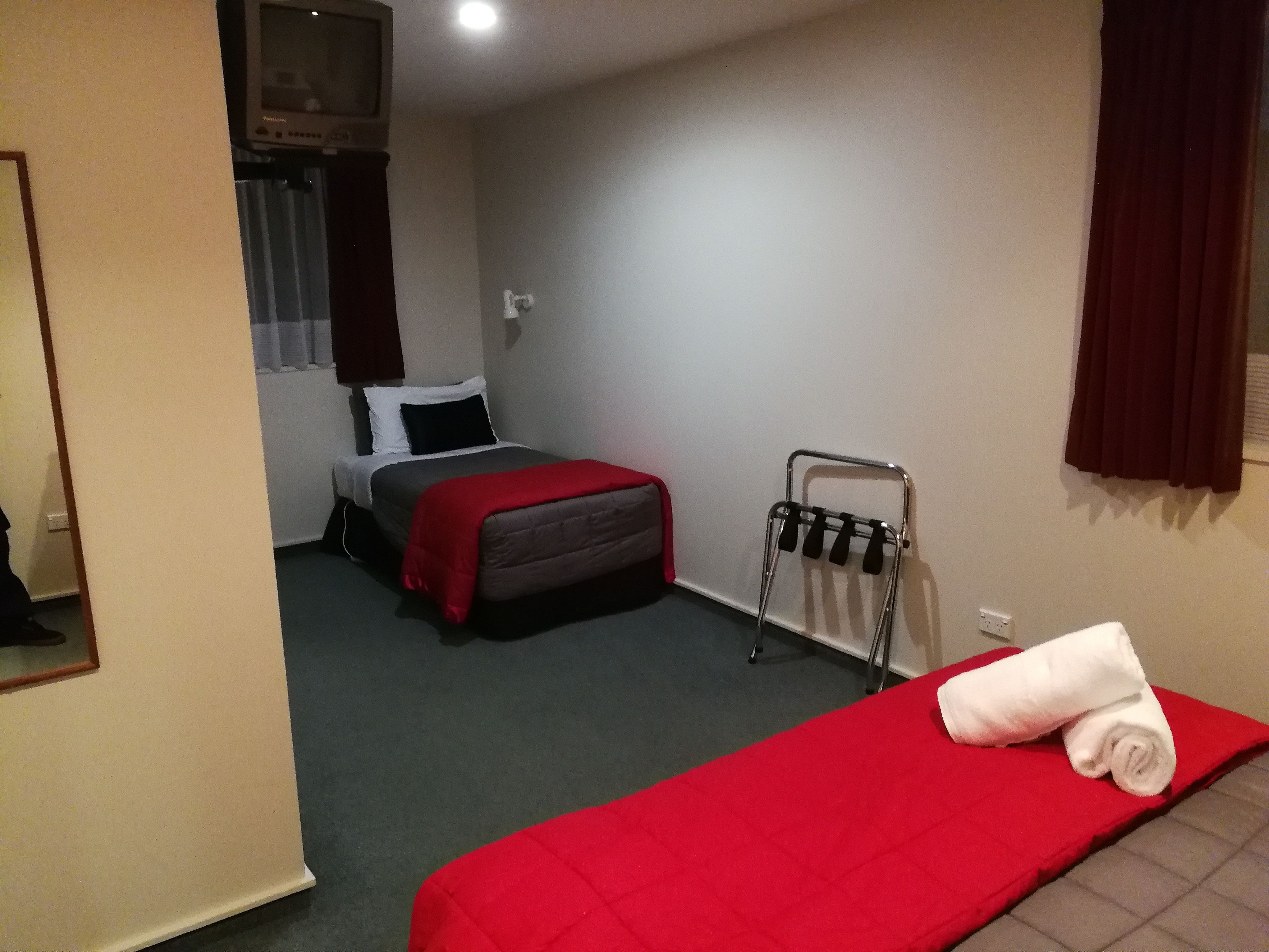 Habitació d'hotel amb un llit individual, una taula petita amb estovatge vermell, un suport de tovalloles fixat a la paret i un televisor penjat a la pared.