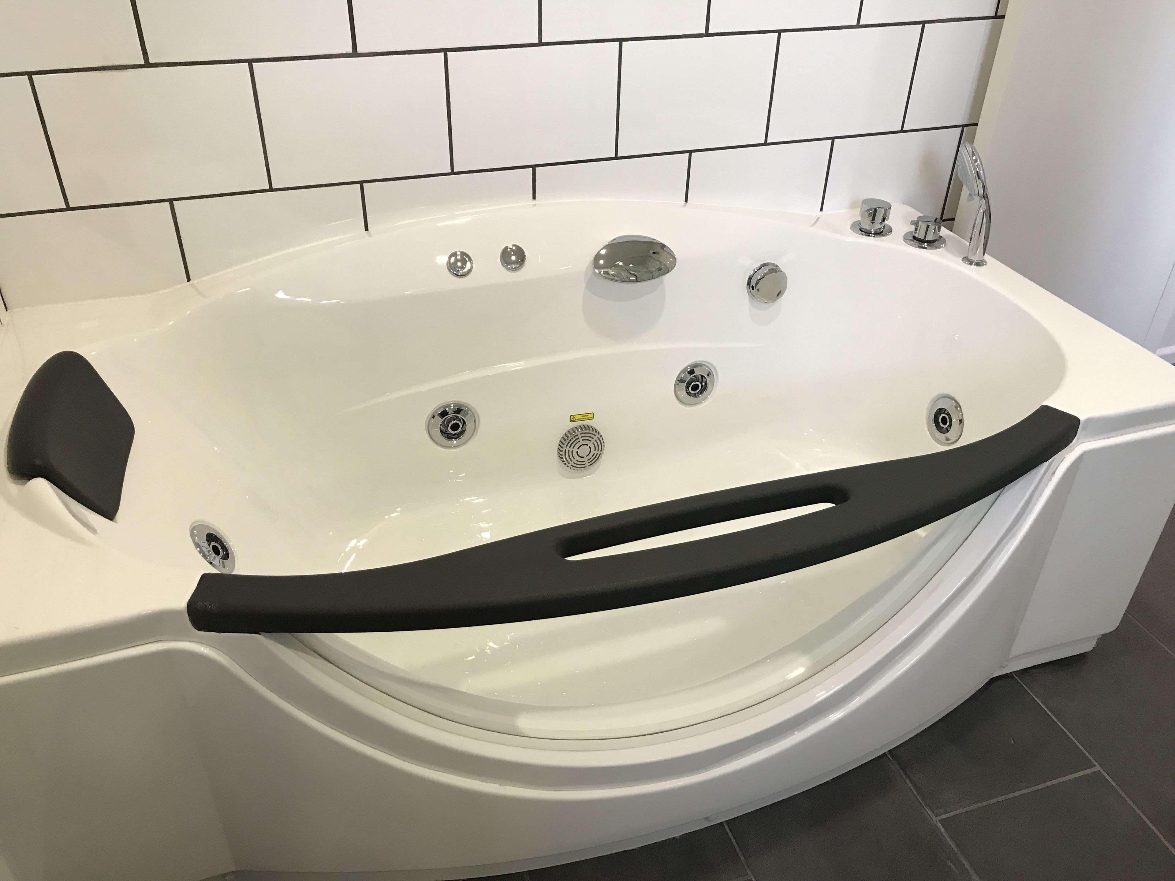 Una bañera blanca con una barra de seguridad negra, grifería de cromo y una pared con azulejos al fondo.