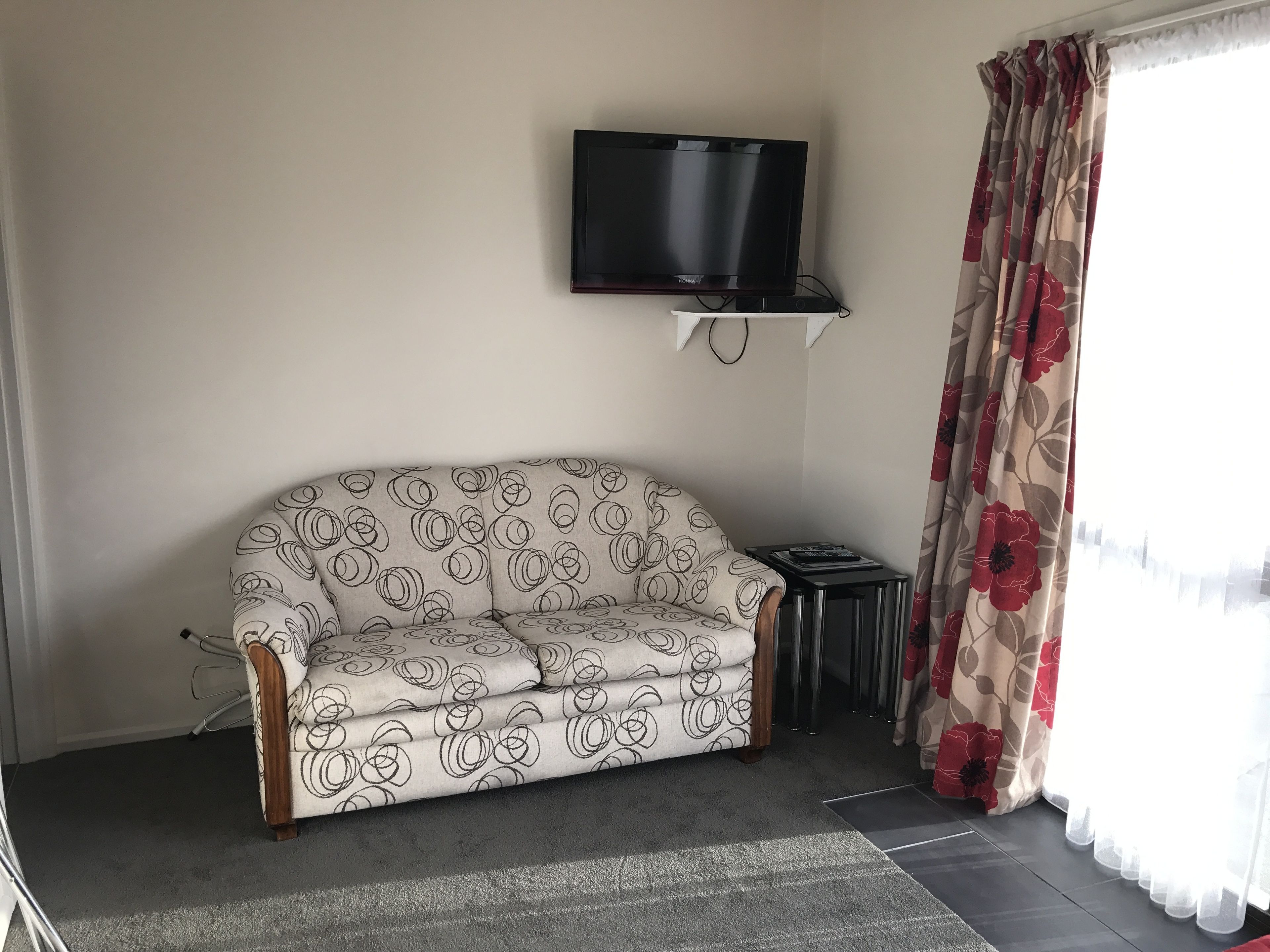 A living room with a sofa, a television mounted on the wall, a small side table, and a window with curtains.