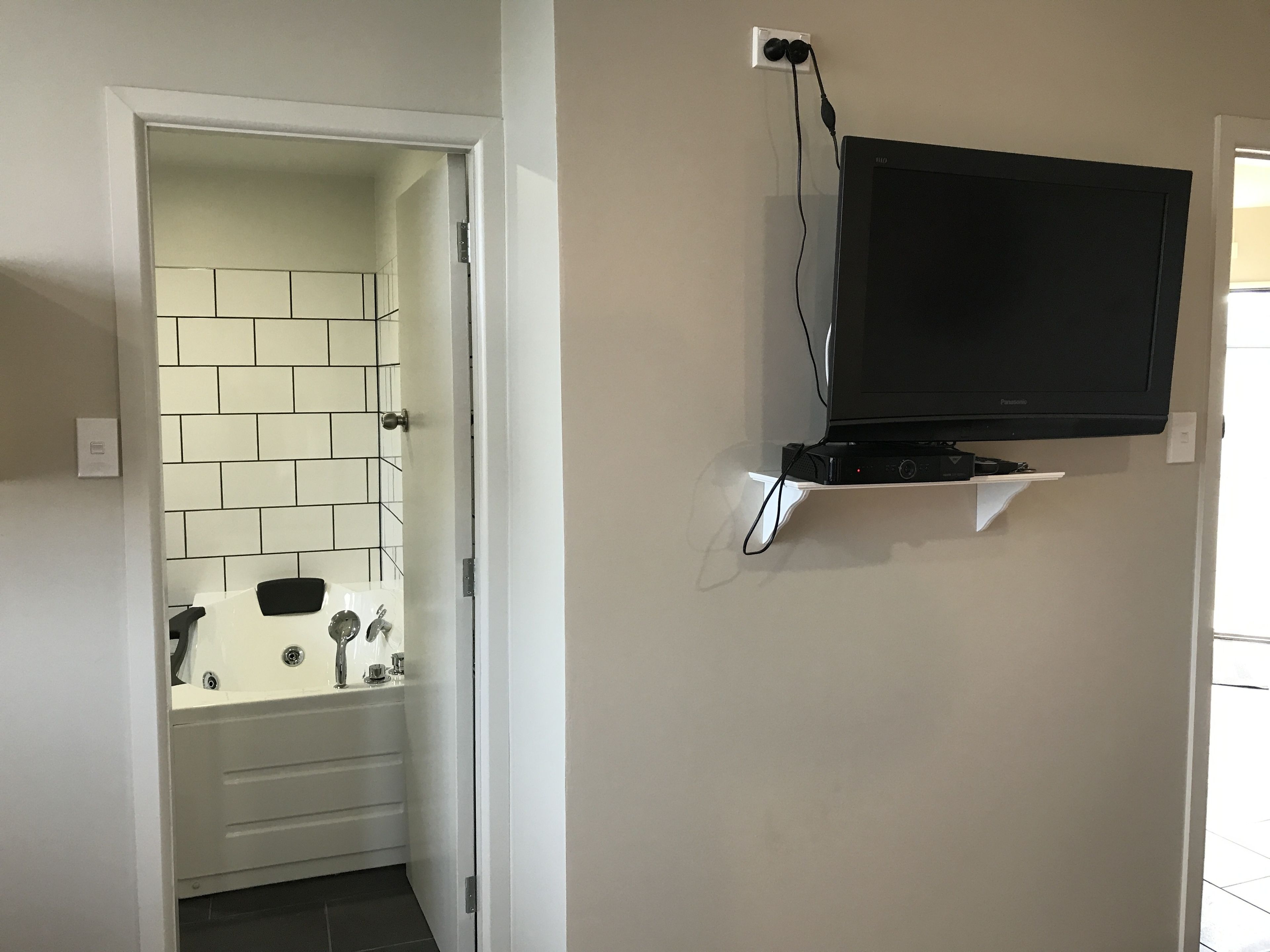 A bathroom with a tiled wall, a bathtub, and a mounted television on a shelf.