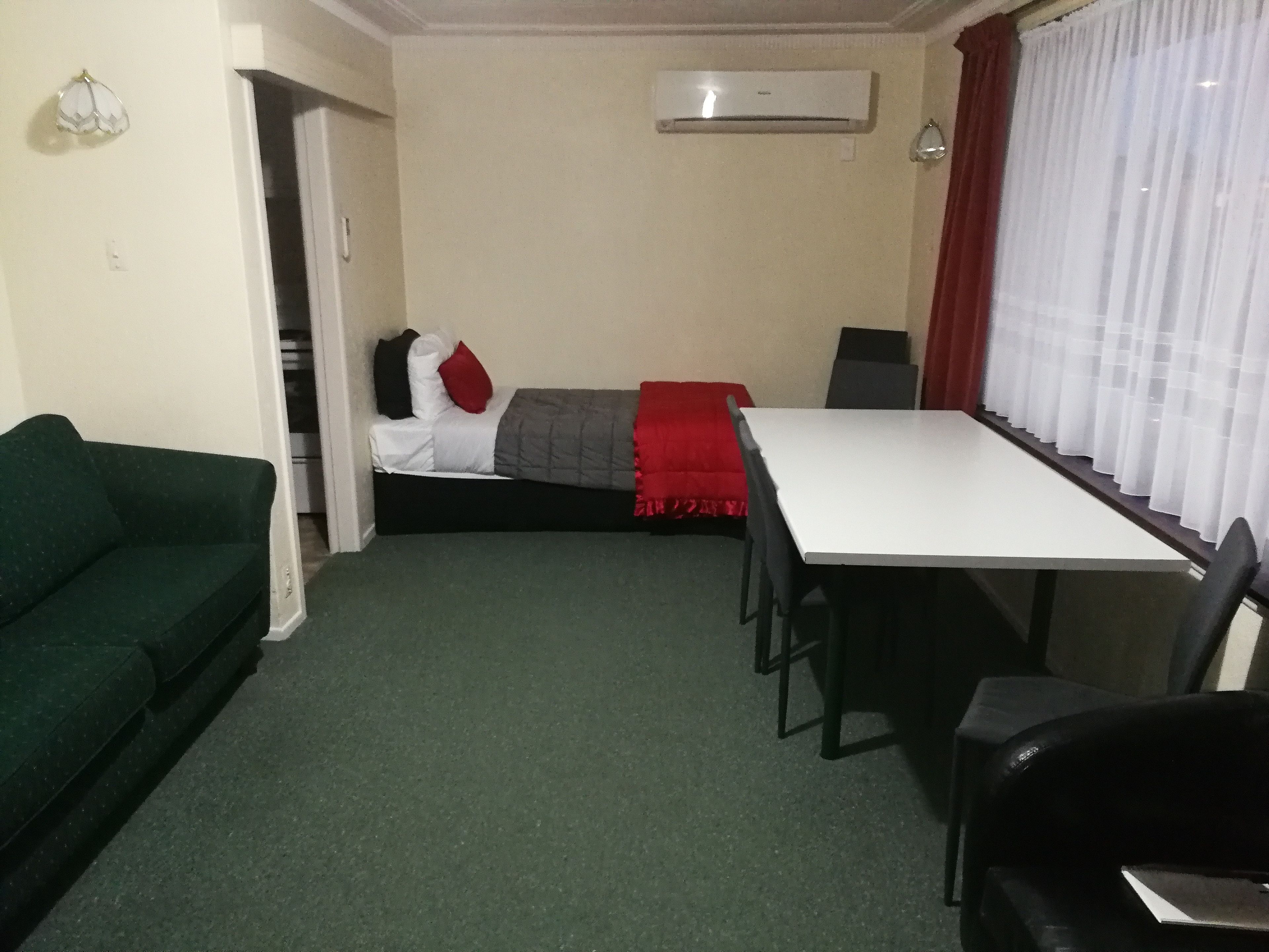 Habitación pequeña y compacta con un sofá verde, una cama blanca con ropa de cama roja y gris, una mesa blanca y sillas negras. Un equipo de aire acondicionado está montado en la pared sobre la cama.