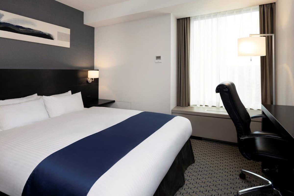 Une chambre d’hôtel avec un grand lit, un bureau avec une chaise, une fenêtre avec des rideaux et une lampe fixée au mur.
