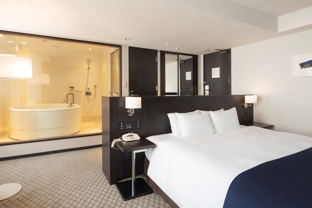 Une chambre d’hôtel moderne avec un grand lit, une table de chevet avec un téléphone, une petite table de nuit et un grand miroir au-dessus du lit.