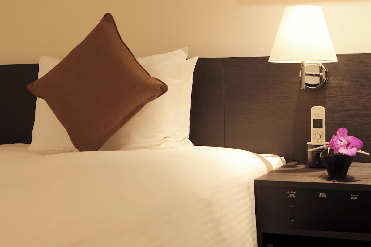 Une chambre d’hôtel avec un lit bien fait, une lampe de chevet, un téléphone et un petit vase contenant une orchidée.