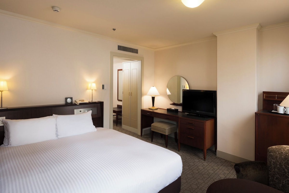 Habitación de hotel con una cama grande, un escritorio con televisor, una silla y una pequeña mesa con una taza.