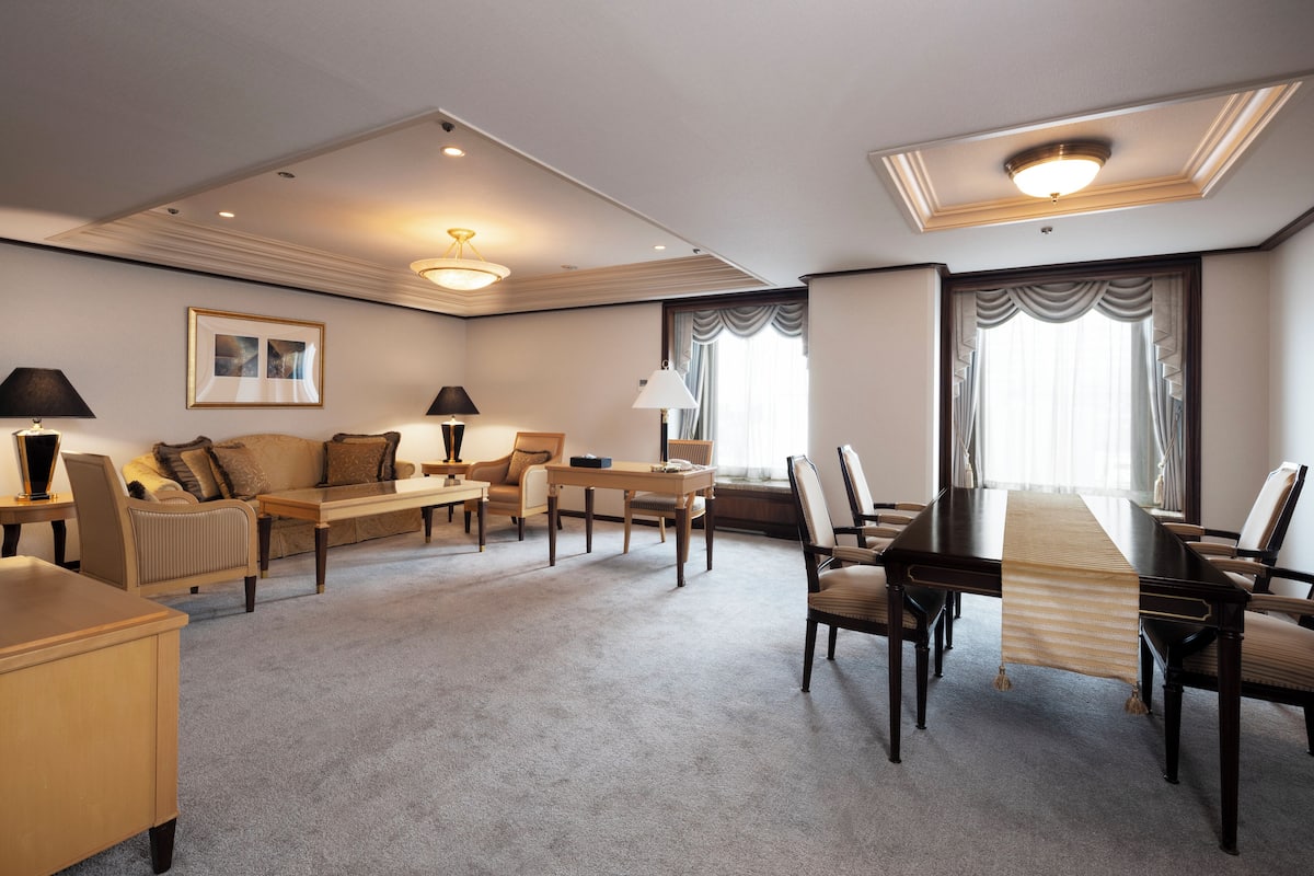 Une chambre d’hôtel dotée d’un grand lit, d’un coin salon avec un canapé et des fauteuils, d’une table à manger avec des chaises, et d’une grande fenêtre avec des rideaux.
