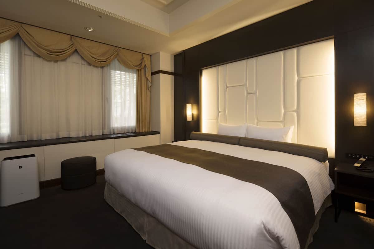 Une chambre d’hôtel avec un grand lit, une table de chevet, une lampe, une fenêtre avec des rideaux et un luminaire fixé au mur.