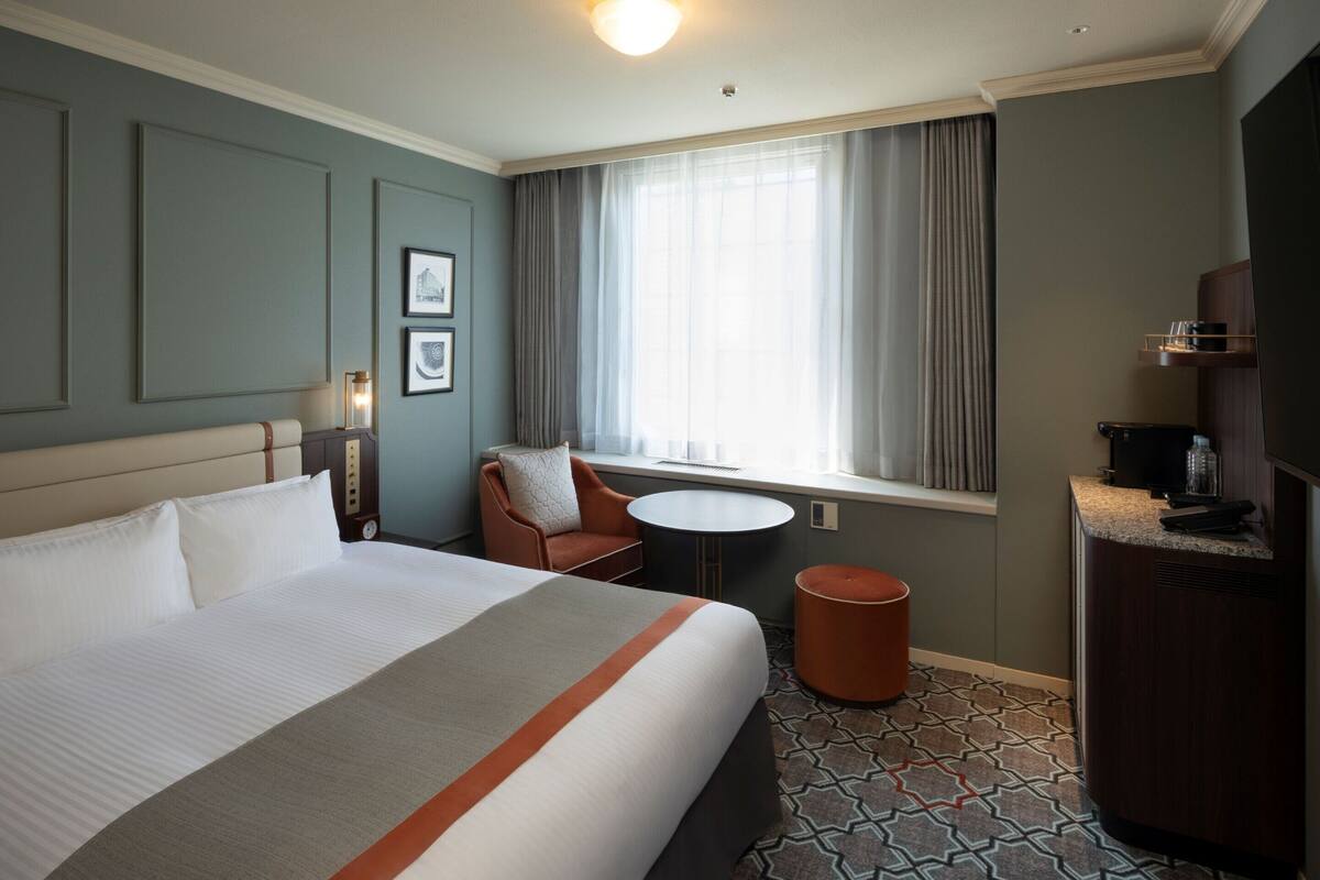 Une chambre d’hôtel avec un grand lit, une petite table ronde, une chaise, un bureau avec un ordinateur et une fenêtre avec des rideaux.