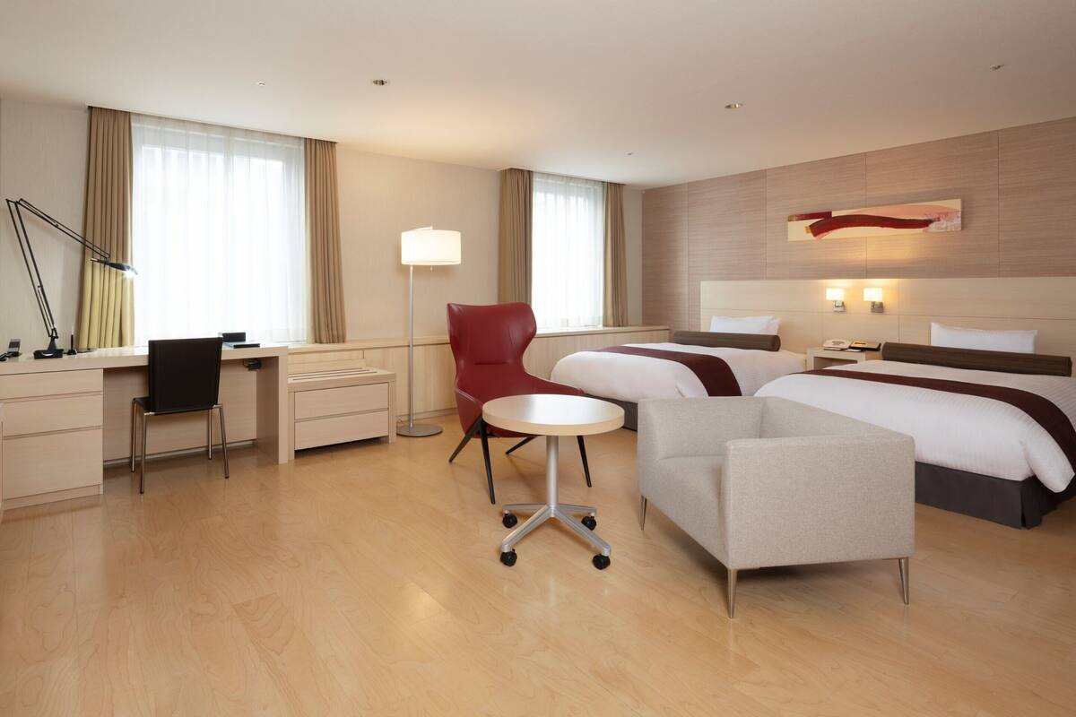 Une chambre d’hôtel moderne, équipée d’un bureau, de deux lits, d’un fauteuil rouge, d’une table ronde et d’une grande fenêtre avec des rideaux.