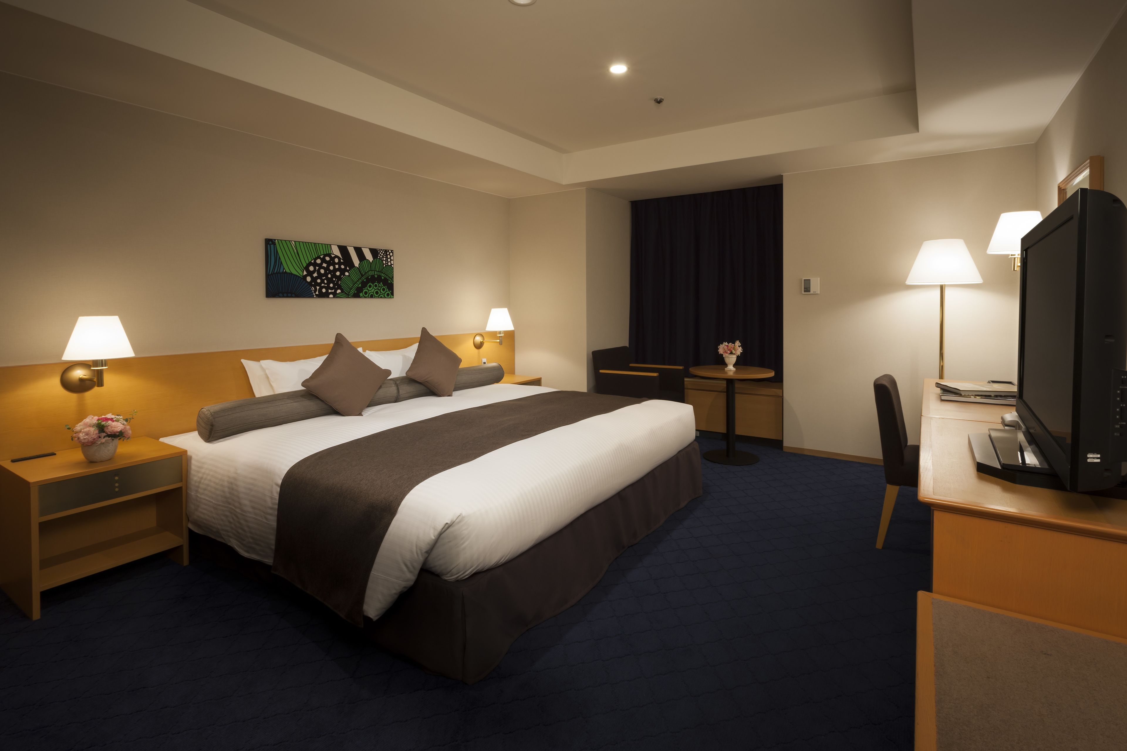 Habitación de hotel con una cama grande, lámparas de noche, un escritorio con televisor y una mesita con lámpara.