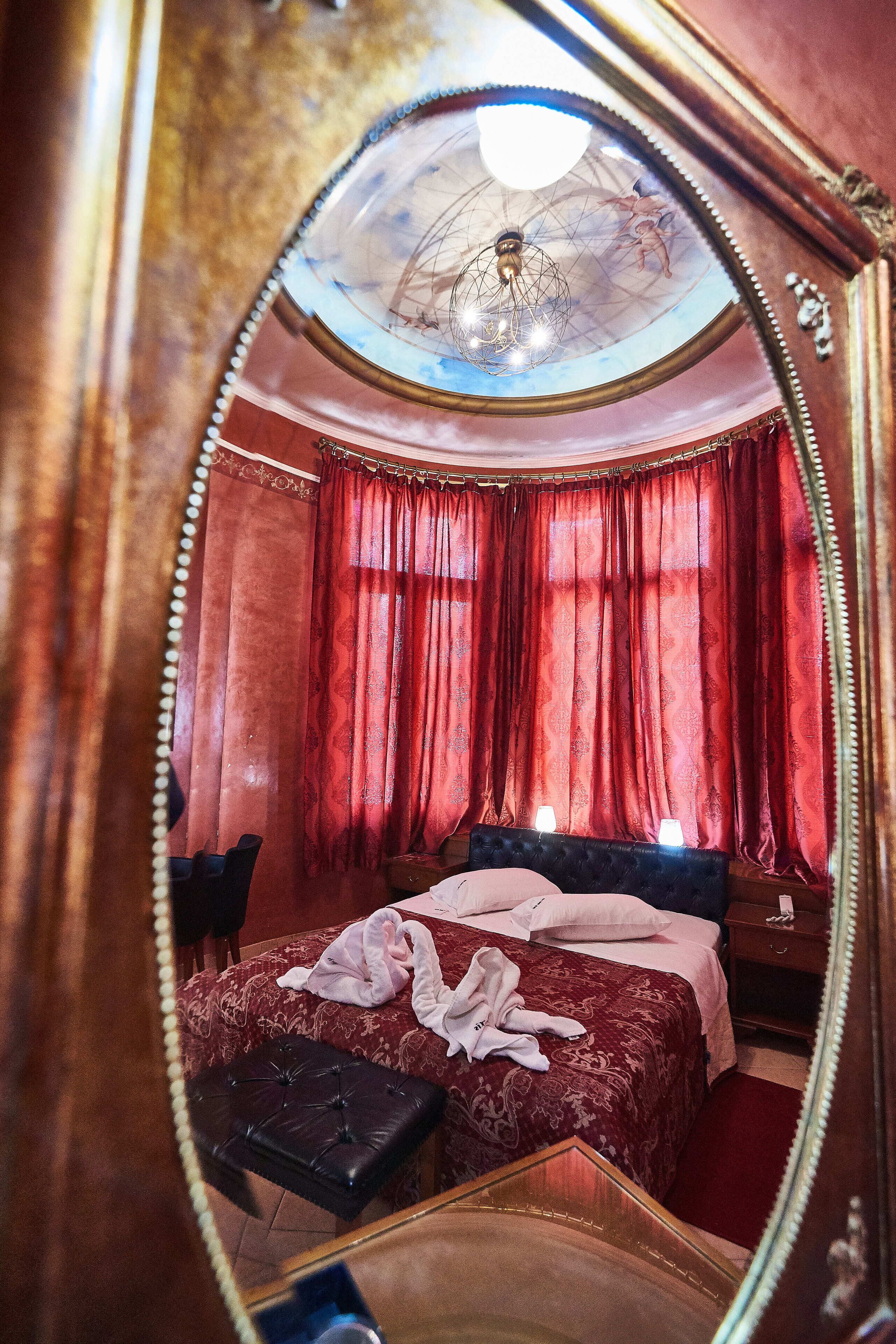 A bedroom with a large mirror, a bed with a red bedspread, a black ottoman, and a chandelier.
