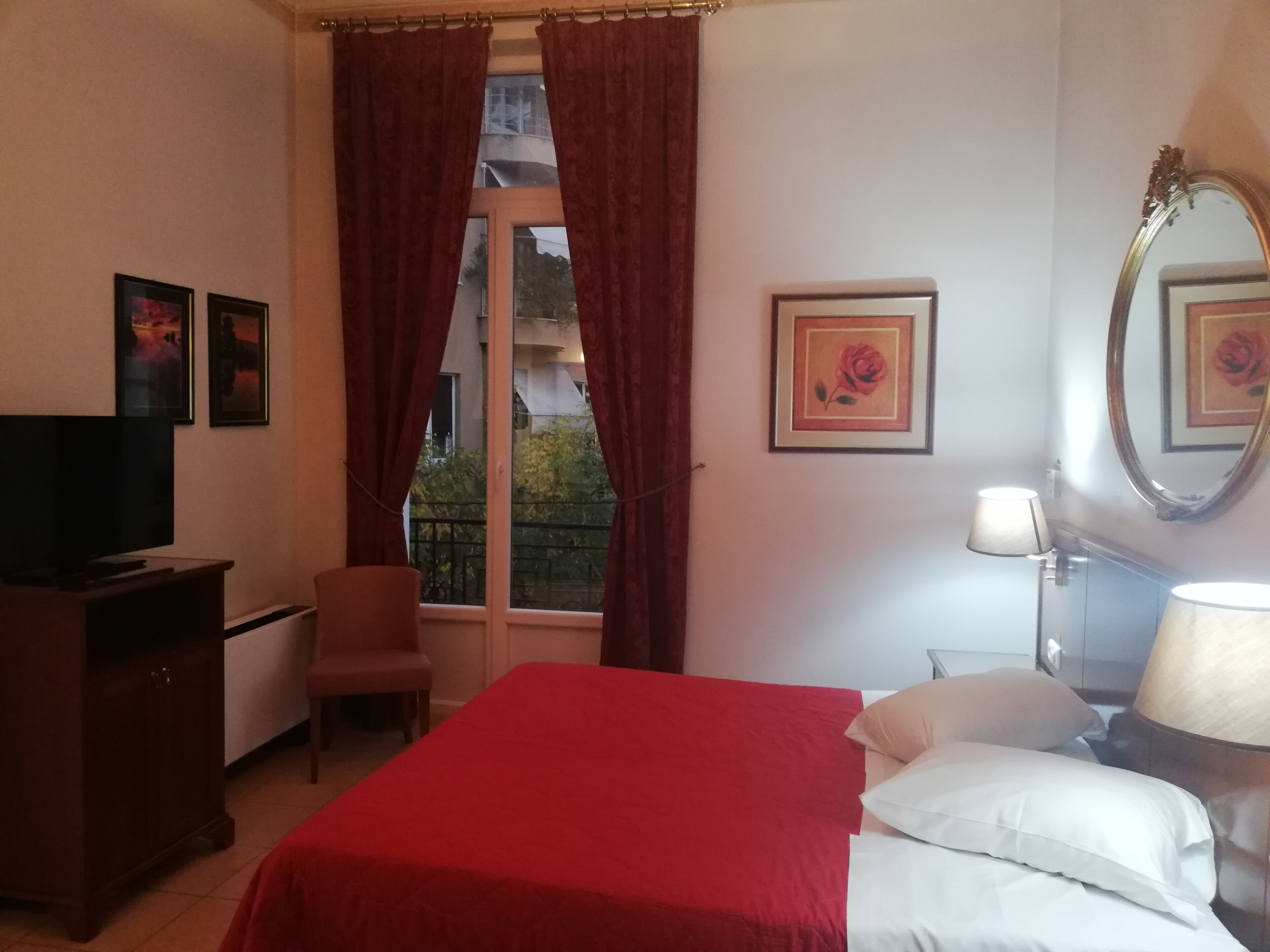 A hotel room with a bed, a desk, a chair, a TV, a window with curtains, a lamp, and framed pictures on the wall.