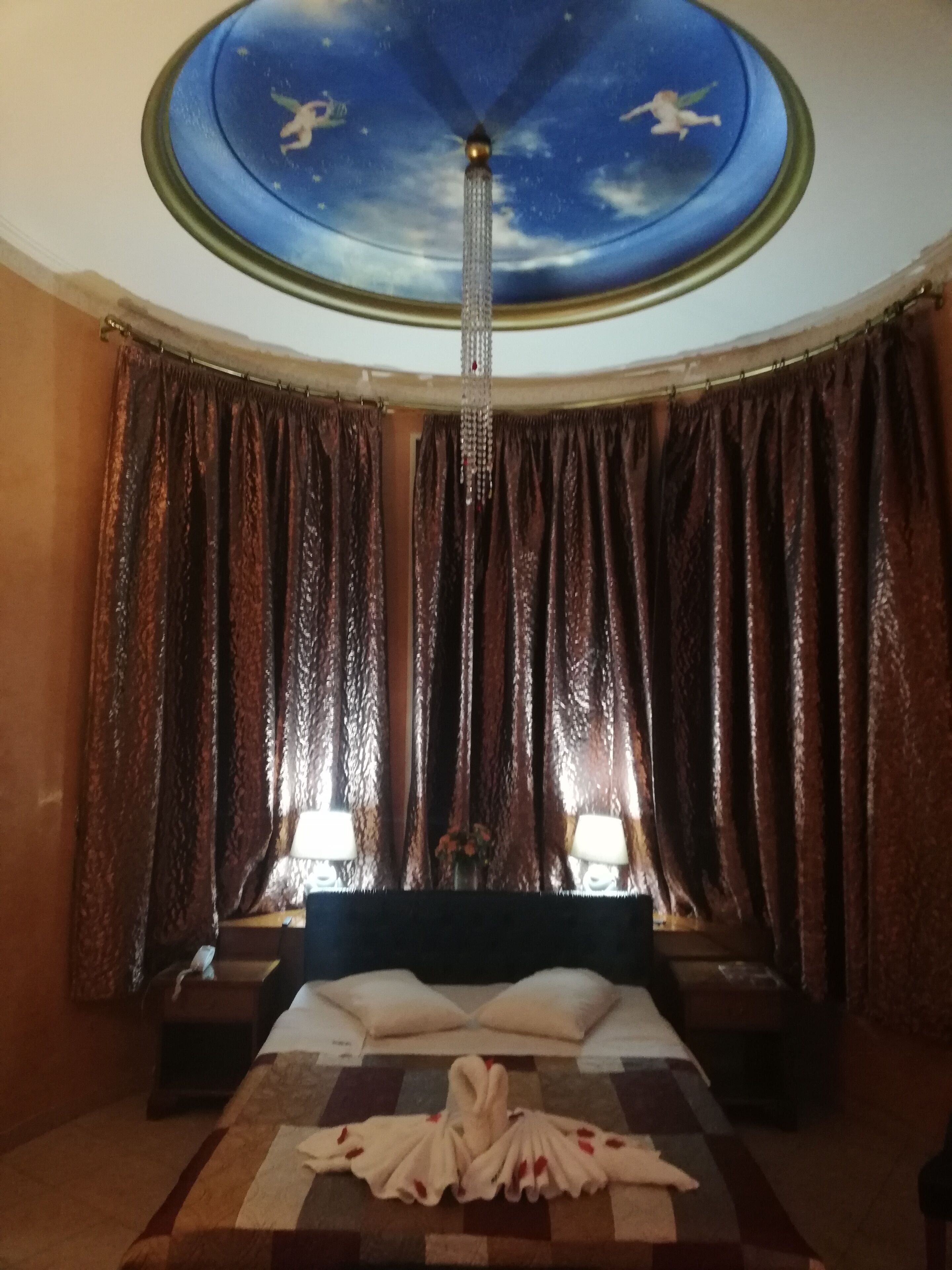 Una habitación de hotel con cama, cortinas, una araña y un mural en el techo.