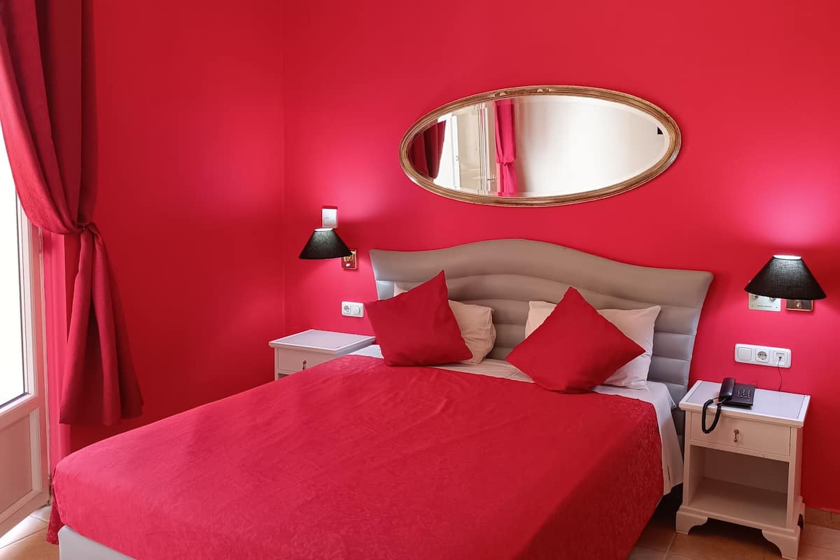 A bedroom with a red wall, a bed with red bedding, a round mirror, two bedside tables with lamps, and a red curtain.