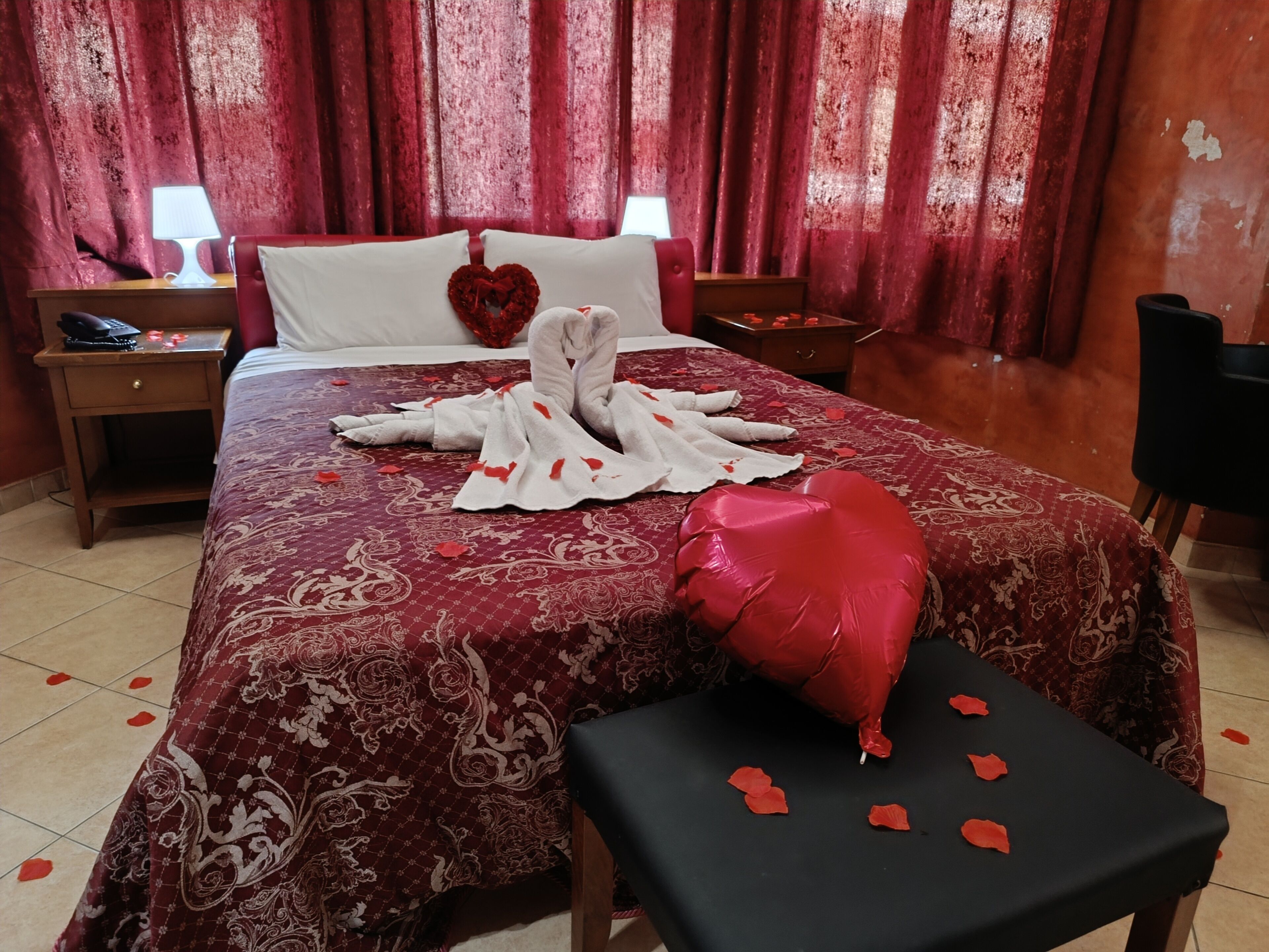 A hotel room with a bed, a nightstand, a red heart-shaped pillow, and a red heart-shaped decoration on the bed.