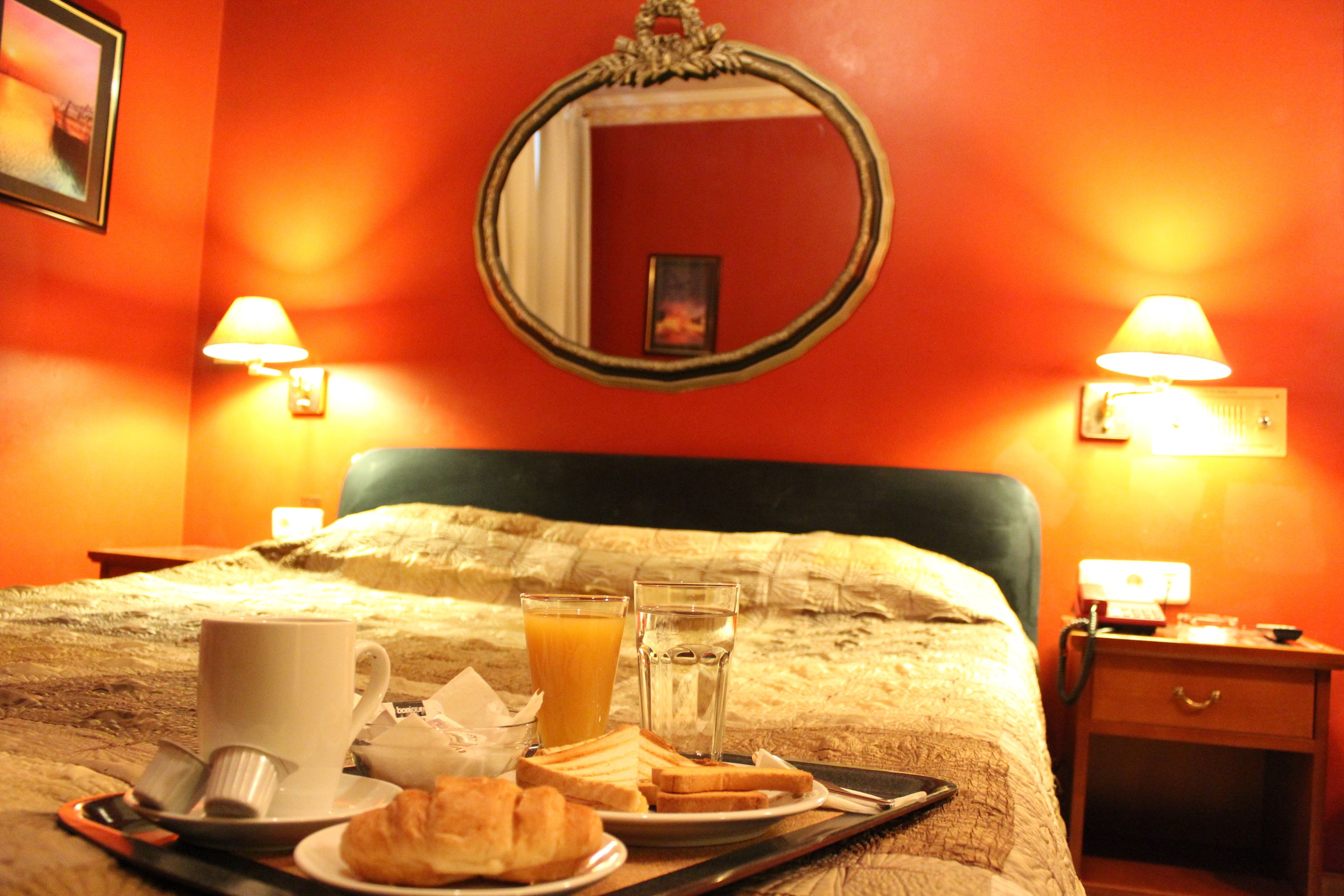 Ein ordentlich gedeckter Frühstückstisch mit Toast, einer Tasse und einem Glas Orangensaft auf einem Bett mit Kopfteil, in einem Zimmer mit orangefarbenen Wänden und zwei Nachttischlampen.