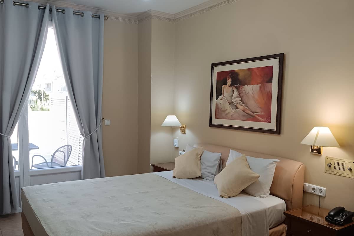 A hotel room with a large bed, two bedside tables with lamps, a ceiling light fixture, a painting on the wall, and a window with curtains.