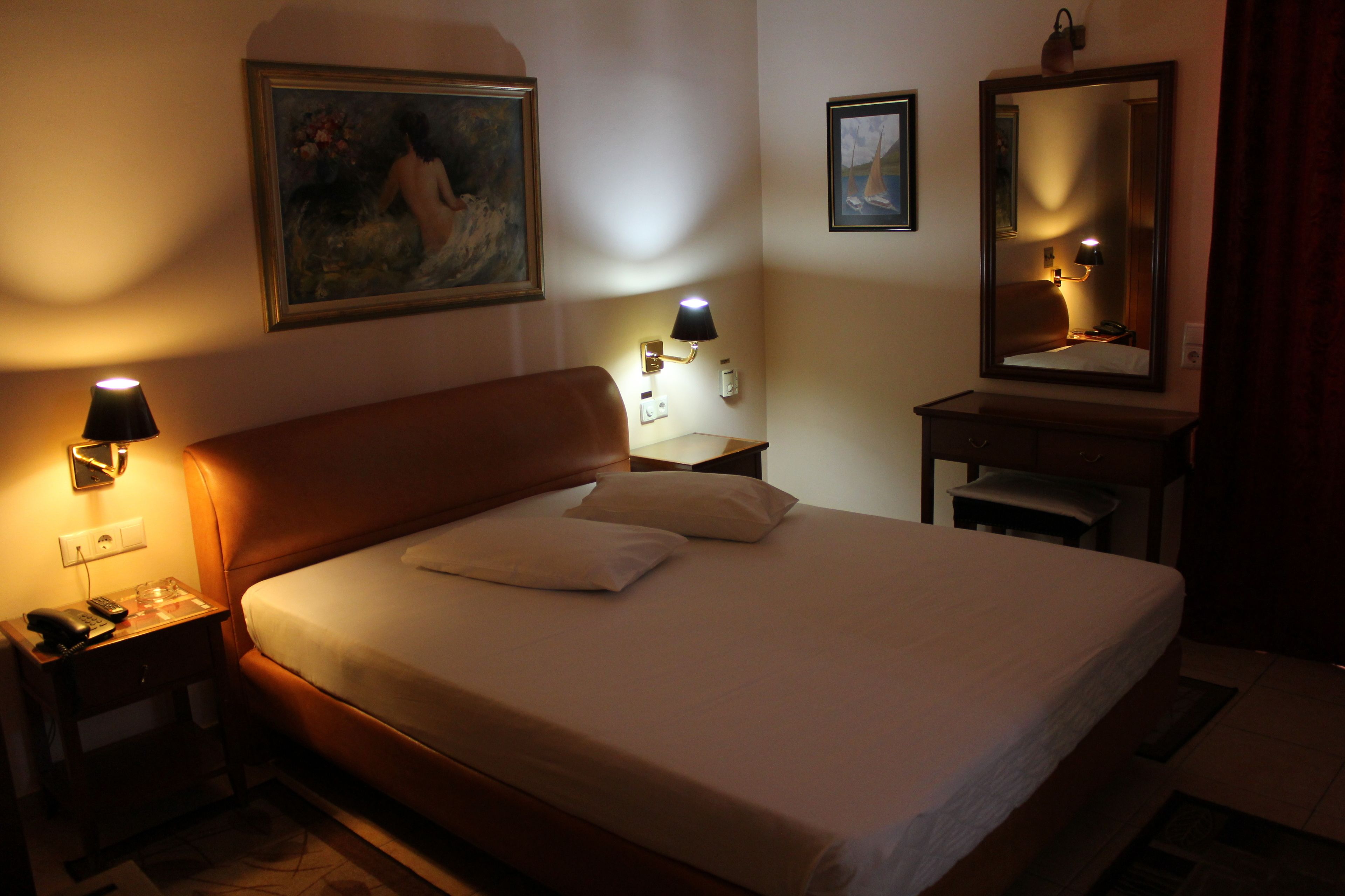 Una habitación de hotel con cama, mesitas de noche, lámpara, cuadro, espejo y escritorio.